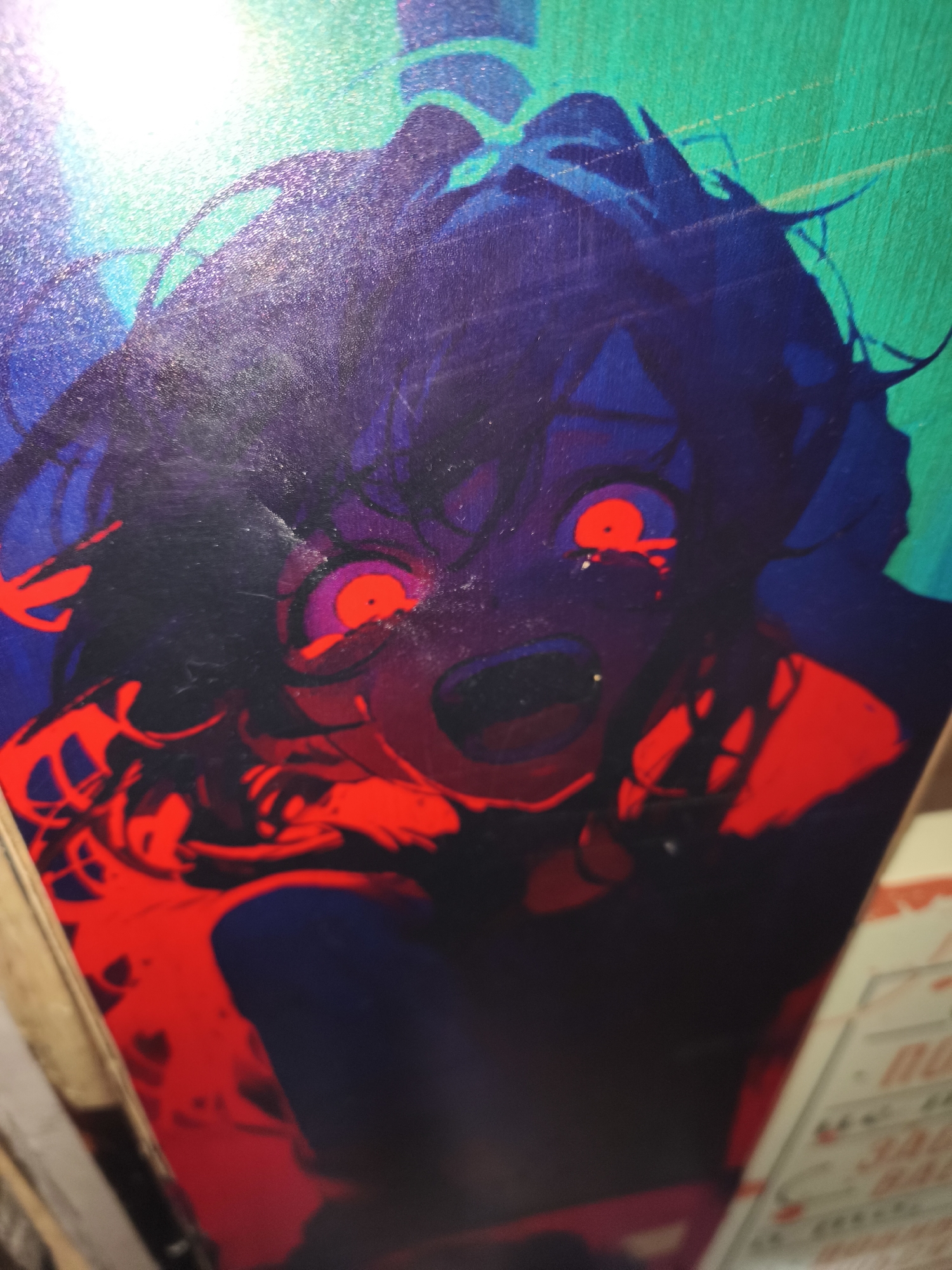 The media file shows a close-up of a graphic design, likely an illustration or a print on an object. The central subject is a stylized anime-esque character with wide, glowing red eyes and an open mouth in a scream or gasp. The character's hair is dark and appears to be in disarray, with wisps extending outwards. The color palette is dominated by dark purples, blues, and vibrant reds, creating a dramatic and possibly unsettling mood. The character's skin appears pale or purplish. The background transitions from a dark blue on the left to a bright, almost neon green on the right, with some lighter, shimmering elements that suggest a glossy or metallic surface, or possibly a light source reflecting off the image.
The overall impression is one of intense emotion, likely fear, shock, or rage, conveyed through the exaggerated facial features and the stark color contrast. There are no clear indications of a specific time of day or weather conditions, as the image is an artwork. Location cues are absent within the artwork itself.
In the lower right corner, partially visible, is a section of what appears to be packaging or a label with some text. The text is blurry and difficult to read, but some characters and block lettering are discernible in white and orange. There are also some faint lines and patterns that suggest a printed design. Given the context of Kyiv, Ukraine, this could be related to a product or artwork found in that location.