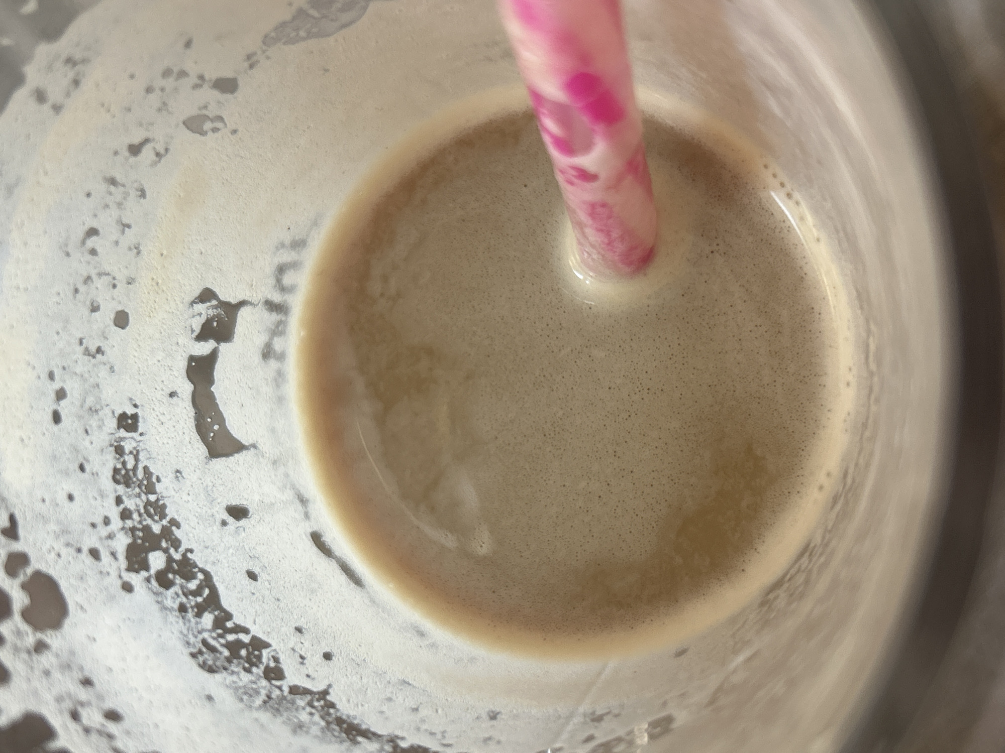 The image is a close-up, overhead shot of a glass containing a foamy, light brown beverage, possibly coffee or a smoothie. A pink and white striped straw is visible, inserted into the drink. The glass appears to be partially filled, with a frothy rim around the edges. The background is indistinct. There are no discernible Plovdiv, Bulgaria landmarks or environmental cues in this image.