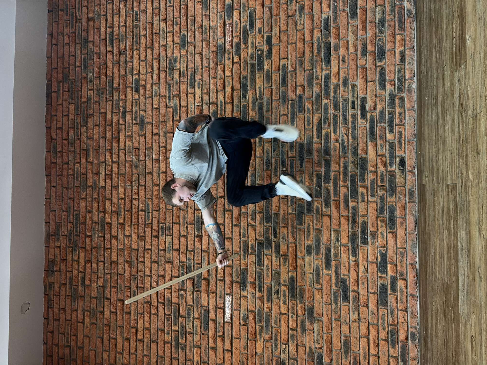 The image displays a man in an dynamic, acrobatic-like pose, appearing to balance on a vertical brick wall. The photograph is rotated 90 degrees clockwise, creating the illusion of the man defying gravity against the wall. He is positioned centrally, wearing a light grey t-shirt, dark pants, and white athletic shoes. Visible tattoos adorn both of his arms. His left hand is braced against the textured brick surface, while his right hand firmly grips a long, slender wooden stick extending downwards. His body is bent, with his legs also against the wall, suggesting a feat of strength or balance. The man has short hair and a beard, and his expression is one of focus or concentration.
The primary background is a prominent brick wall, characterized by an irregular pattern of reddish-brown and dark grey/black bricks arranged vertically. To the right of the brick wall, a section of light brown wooden flooring or paneling is visible, also running vertically. On the far left, a plain white wall forms a corner with the brickwork. The setting appears to be indoors, possibly a studio or training space in Chaikovsky, Russia, with even, artificial lighting. Faint, illegible text is discernible on the man's t-shirt.