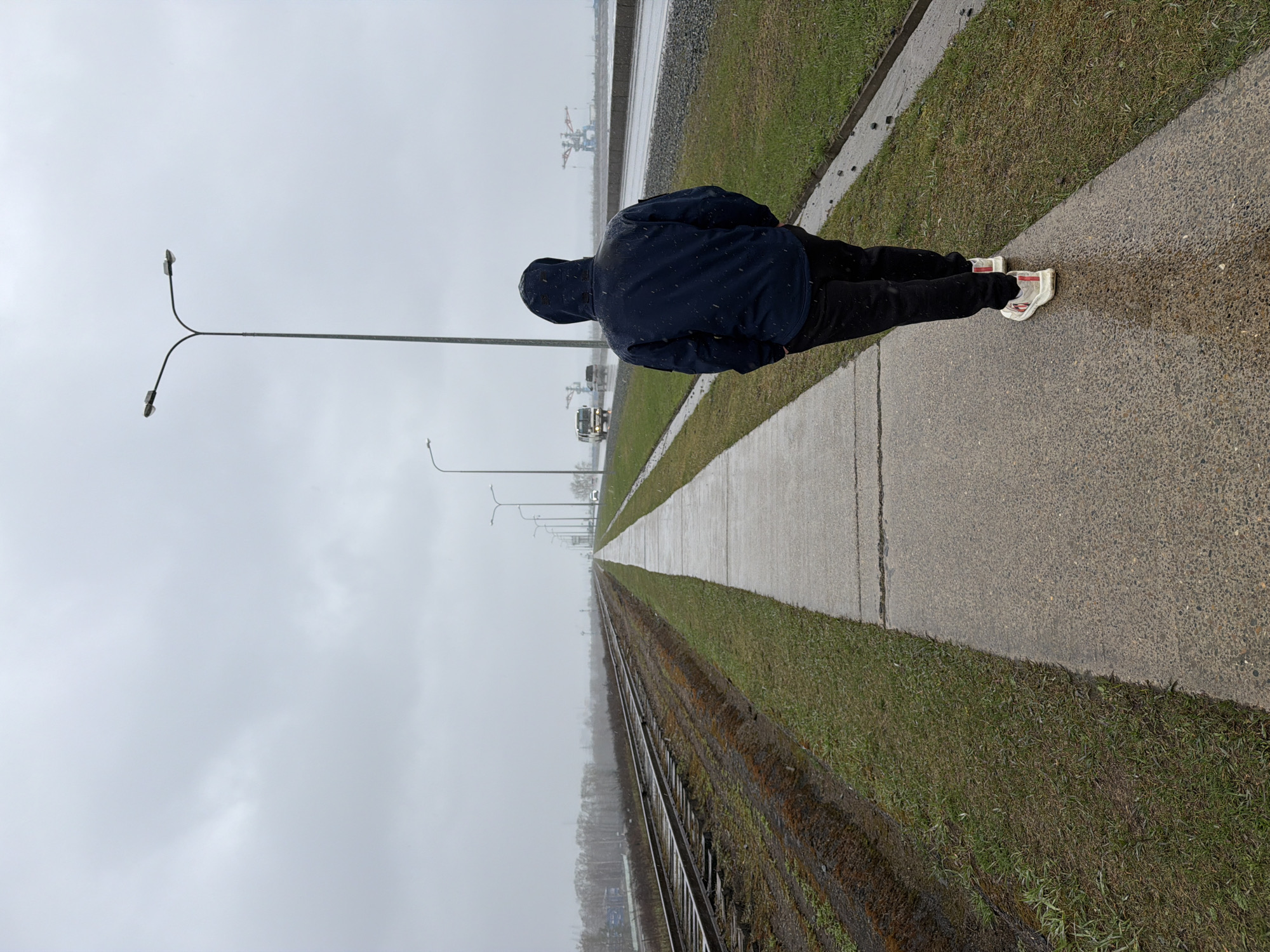 The image captures a person from behind, walking or standing on a paved path under a dull, overcast sky. The individual wears a dark blue hooded jacket, dark pants, and white sneakers with red accents. Small white specks, indicative of light snow or rain, are visible on their jacket and the wet ground.
The paved path runs parallel to a strip of green grass, which in turn borders railway tracks on the left side of the frame. Both the path and the tracks extend far into the misty distance, creating a strong sense of linear perspective. Along the path, multiple street lights with dual lamps also recede into the background.
In the hazy distance, beyond the railway tracks, industrial structures resembling cranes are visible, along with what appear to be distant vehicles (possibly trucks), suggesting an industrial area, likely a port or transport hub. The sky is uniformly grey and overcast, and the ground surfaces appear wet from recent or ongoing precipitation. The overall atmosphere is stark and subdued. There is no visible text in the image. The scene is set in an outer or industrial part of Chaikovsky, Russia.