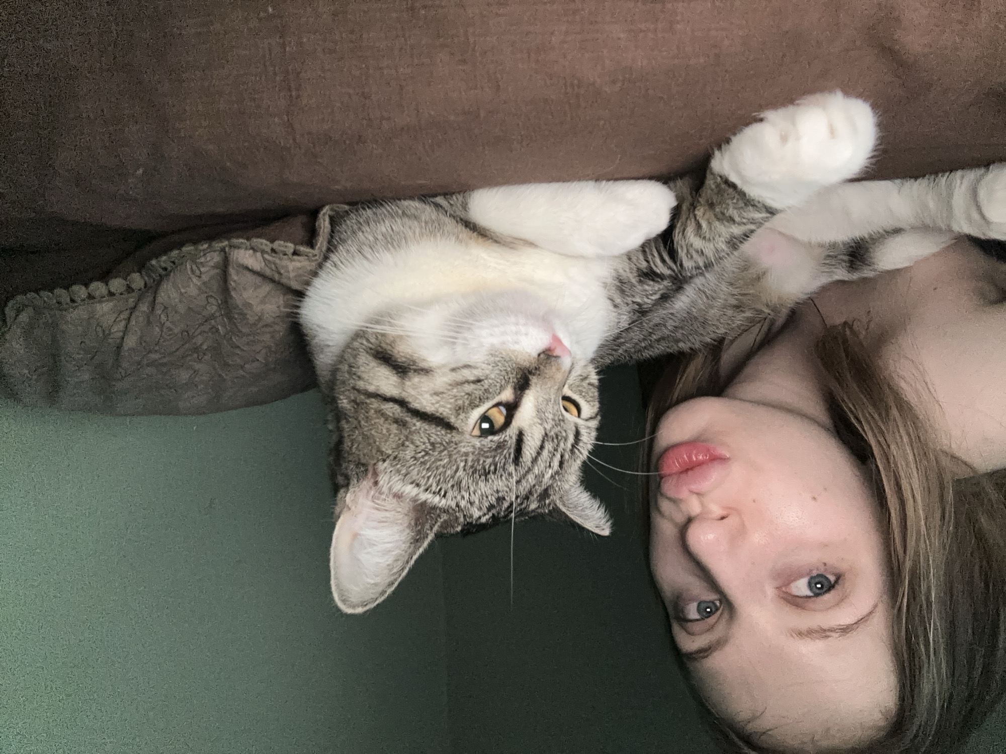 An indoor, close-up scene depicts a female individual and a domestic cat. The female, positioned on the left side of the frame, has light hair and blue eyes, and her lips are pursed in a kissing gesture, directed towards the camera. She appears bare-shouldered. Adjacent to her, occupying the center-right, is a domestic short-haired cat with tabby markings and a white chest, belly, and paws. The cat, possessing amber eyes and a pink nose, is lying down on a dark brown surface and looking towards the left, away from the camera. A dark green wall forms the background, with a textured, dark gray pillow or blanket visible behind the cat's head. A single, thin, light-colored fiber extends vertically in the air between the female's face and the cat's ear. The specific location within Chaikovsky, Russia, cannot be determined from the visual information presented.