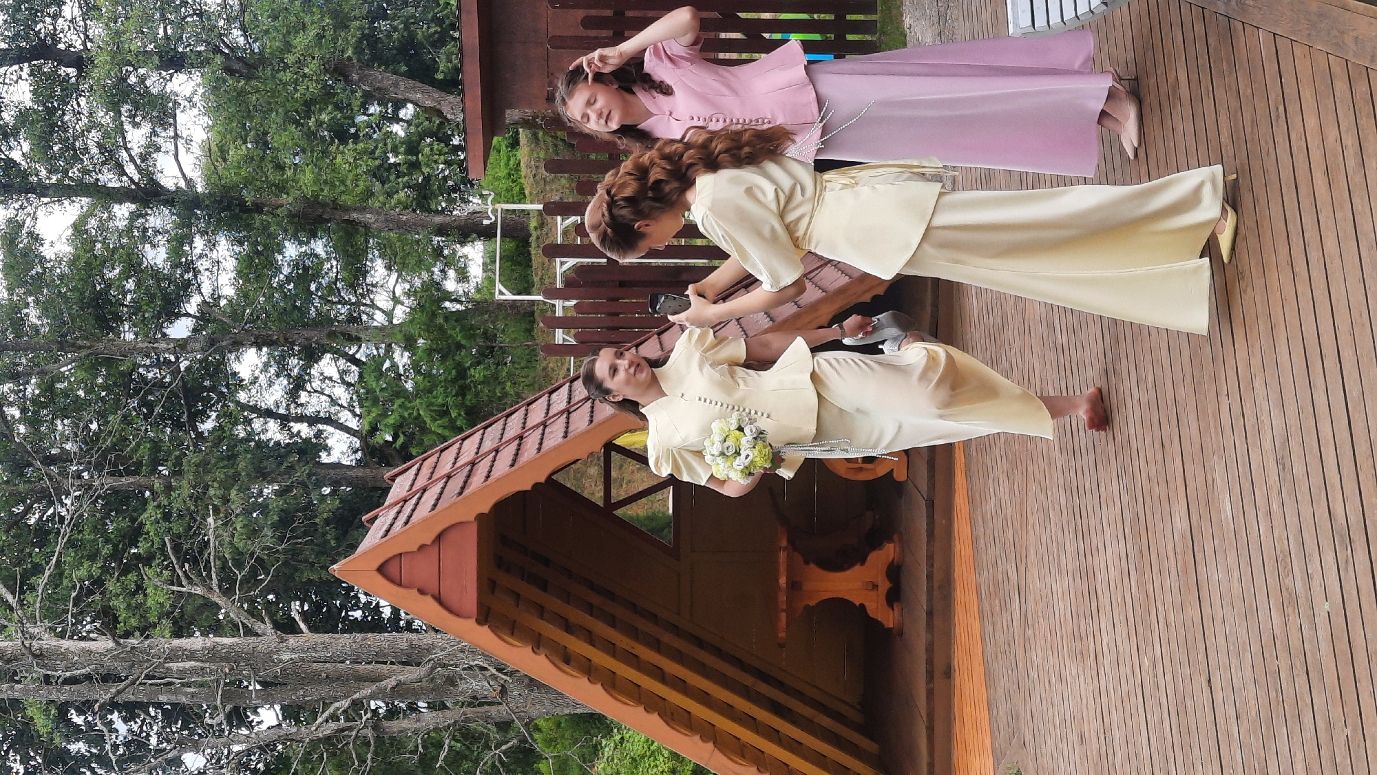 Three young women are gathered on a wooden deck outdoors, near a small, wooden A-frame structure with a reddish-brown, shingled roof. The woman on the left, wearing a pale yellow outfit with a peplum top and long skirt, holds a bouquet of white and green flowers and is barefoot. The woman in the middle, also in a pale yellow outfit consisting of a similar peplum top and long skirt, is taking a photo of the woman on the left with her smartphone. The woman on the right wears a pink, peplum-style top and a long pink skirt, and she is touching her hair. Behind them, a wooden fence is visible. The environment is natural, with trees and foliage surrounding the area. The overall scene suggests a casual outdoor gathering or event.