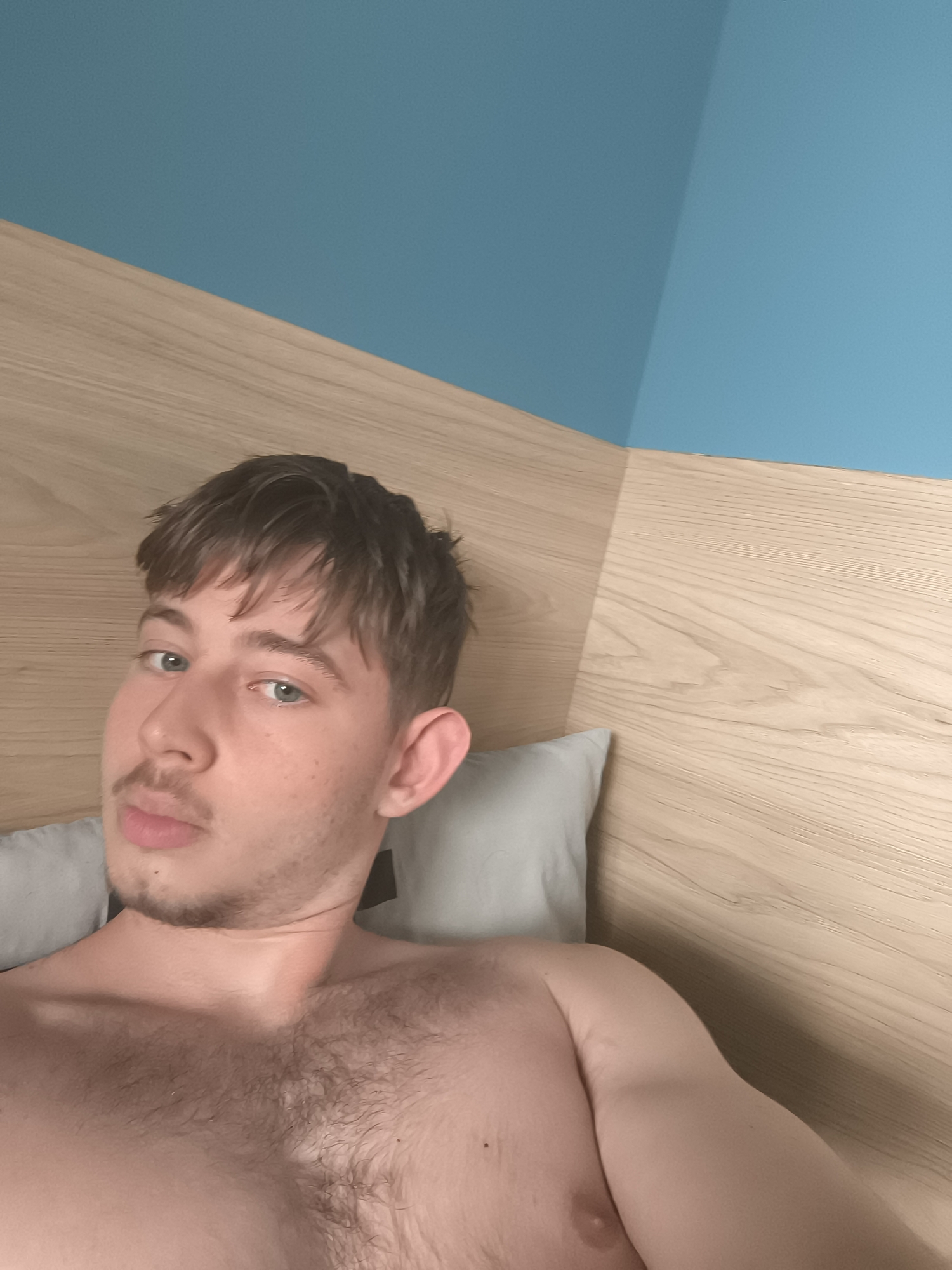 A shirtless, fair-skinned man lies in bed. He has short brown hair with a fringe, a light beard, and blue eyes. He is propped up against gray pillows and a wooden headboard. The wall behind the headboard is painted blue in the top corner. The location is unidentifiable as being within Sofia, Bulgaria, based solely on the visual information available in the image.
