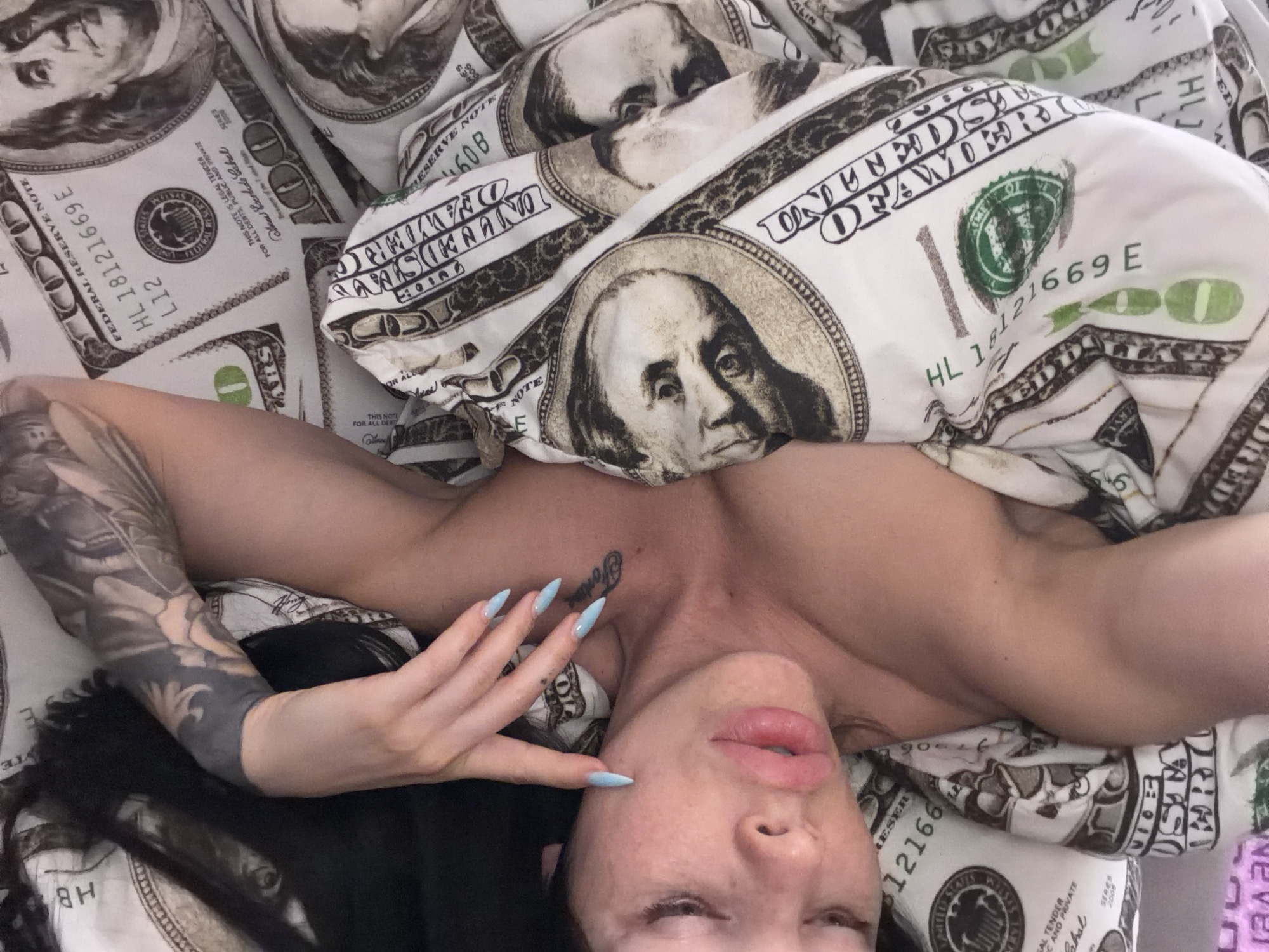 A woman is lying on a bed with sheets patterned with US $100 bills. Her upper body is visible, and she is covered by the patterned sheets. She has dark hair and is wearing light blue acrylic nails. A tattoo is visible on her chest, and another sleeve tattoo is visible on her left arm. The image does not contain information indicating the location of Ufa, Russia.