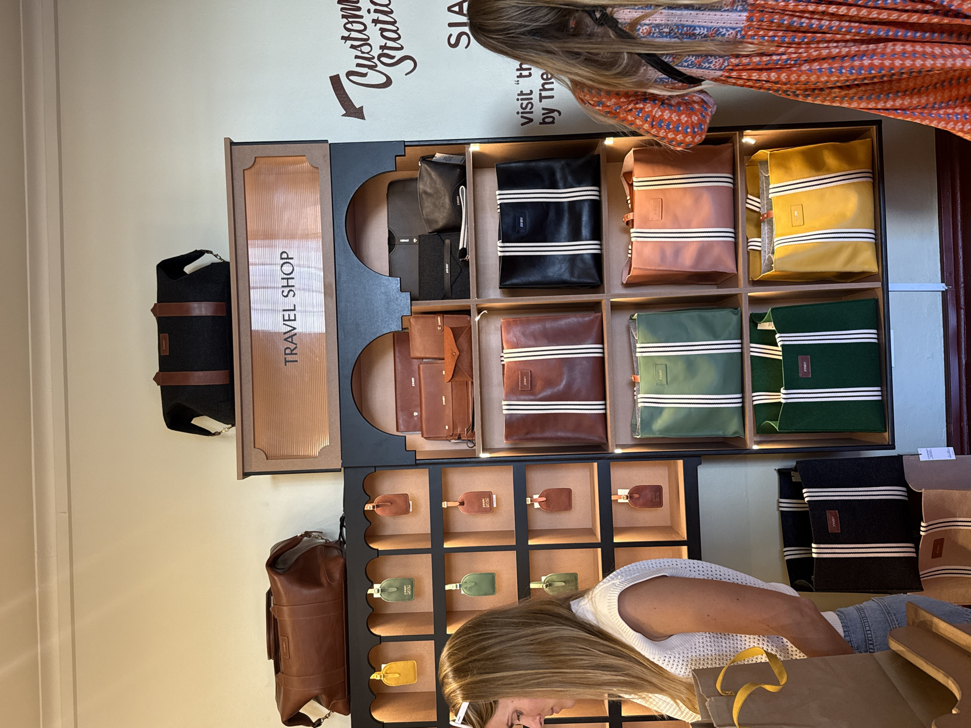 The image depicts the interior of a travel shop in an unknown city. A woman in a white sweater and jeans is looking at travel accessories, specifically luggage tags, displayed in a well-lit wooden shelving unit. The shelving unit also contains various travel bags in different colors such as black, brown, green, pink and yellow. Another woman, wearing an orange patterned dress, is also present looking at the items. The shelving unit also has a section labeled "TRAVEL SHOP". A sign on the wall points to a "Customer Station" with the message "Visit 'this' by The...". Additional travel bags are seen hanging outside the shelves.