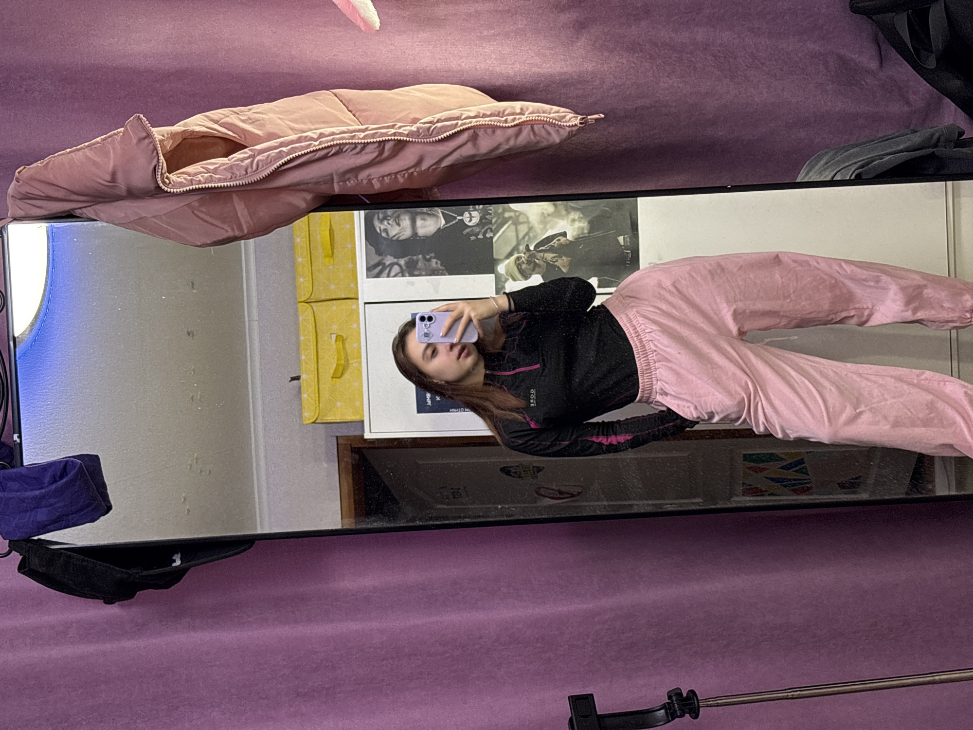 The image shows a mirror selfie of a woman. She is holding a lavender-colored smartphone and wearing a black long-sleeve top with pink accents and pink sweatpants. The mirror reflects part of a room with yellow storage boxes, a framed photograph, and various objects including a pink puffy jacket, a grey bag, and a black hat. The background is a solid light purple color. The location is not discernible from this image.