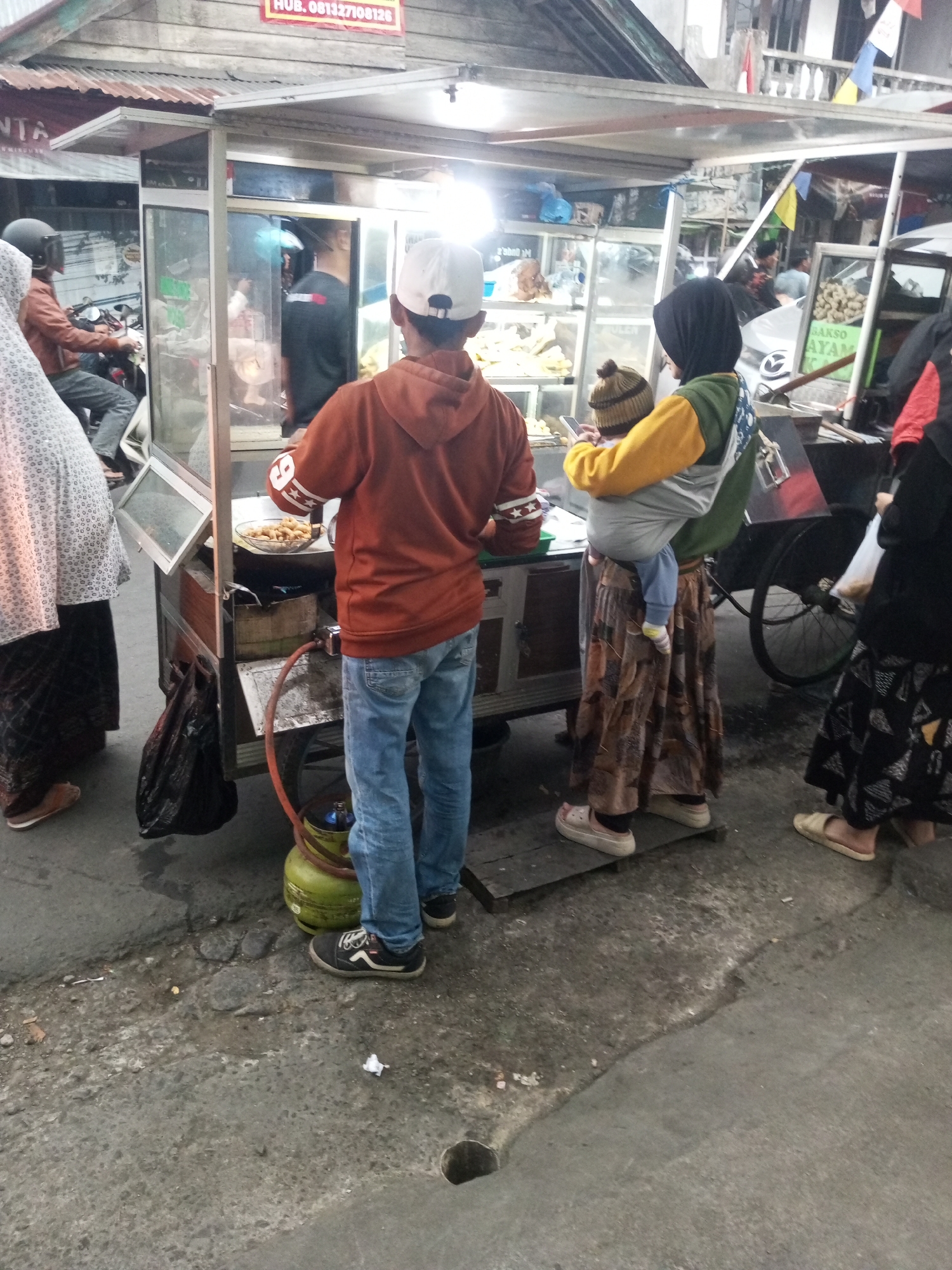 The image depicts a street scene in Kalibeber, Indonesia, near a food cart. In the foreground, a young person wearing an orange hooded sweatshirt and blue jeans stands with their back to the camera, facing the food cart. To the left, another person in a patterned headscarf and long skirt is partially visible. A woman in the middle ground, wearing a dark headscarf, a yellow and green long-sleeved top, and a patterned skirt, is carrying a baby wrapped in a grey carrier. The baby, wearing a knitted hat, faces the camera. The food cart is illuminated by bright lights and displays various food items. A gas cylinder is visible at the base of the cart. In the background, other people and a motorcycle are present. The ground is paved with concrete, and debris is scattered on the surface. The overall atmosphere appears to be that of a bustling street market or food stall area.
