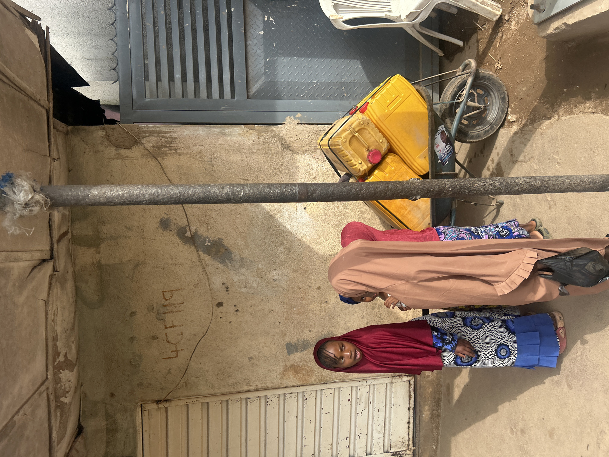 In Jos, Nigeria, three young girls are standing in front of a weathered, light-colored building. The girl on the left, wearing a red headscarf and a blue and white patterned dress with a blue pleated hem, looks to her left with her mouth slightly open. In the center, a girl in a light brown headscarf is partially obscured, facing away from the camera. To her right, another girl, wearing a red headscarf and a pink and blue patterned garment, is partially visible.
Beside them, a rustic metal pole stands vertically. Behind the girls, a yellow wheelbarrow is laden with two large yellow plastic jerrycans, each with a red cap. The wheelbarrow has a metal frame and a single wheel.
The building wall behind them is textured and shows signs of age, with some graffiti that appears to read "4CF119". To the left of the building, a light-colored, corrugated metal roll-up door is visible. To the right, a gray metal gate with horizontal slats is partially open, revealing a dark interior. A white plastic chair sits to the right of the gate. The ground is a mixture of concrete and packed dirt.