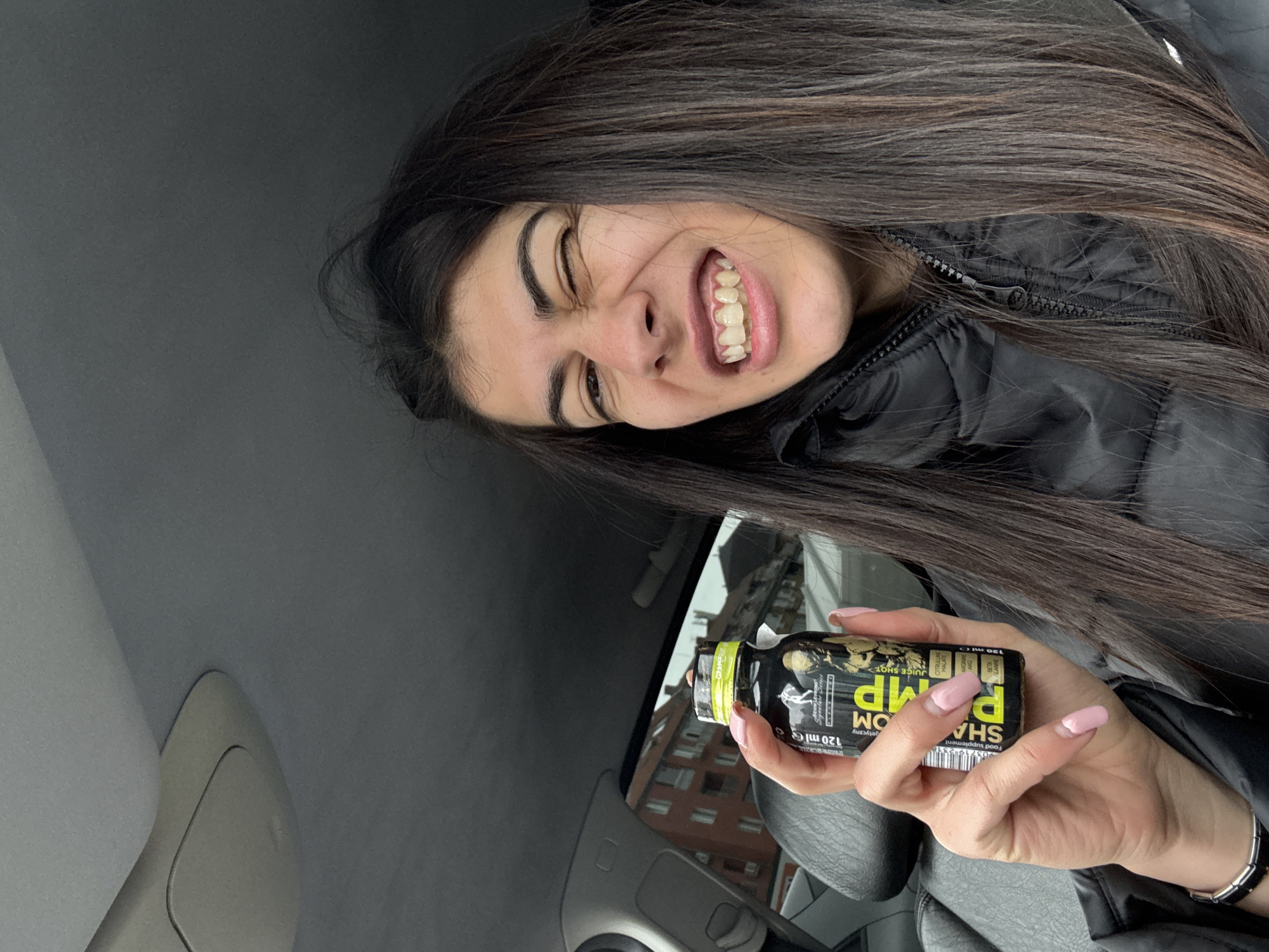A young woman with long, dark hair is holding a small, black and yellow bottle labeled "SHARP PUMP" in her right hand. She is inside a car, possibly a passenger, judging by the dashboard and interior visible. She is smiling broadly and winking one eye. The bottle appears to be a food supplement or energy drink. Through the car window, a multi-story brick building can be seen, suggesting an urban environment. Based on the architecture and the general appearance of the building and surroundings, the location is likely Sofia, Bulgaria. The woman is wearing a black jacket. She has a bracelet on her left wrist.