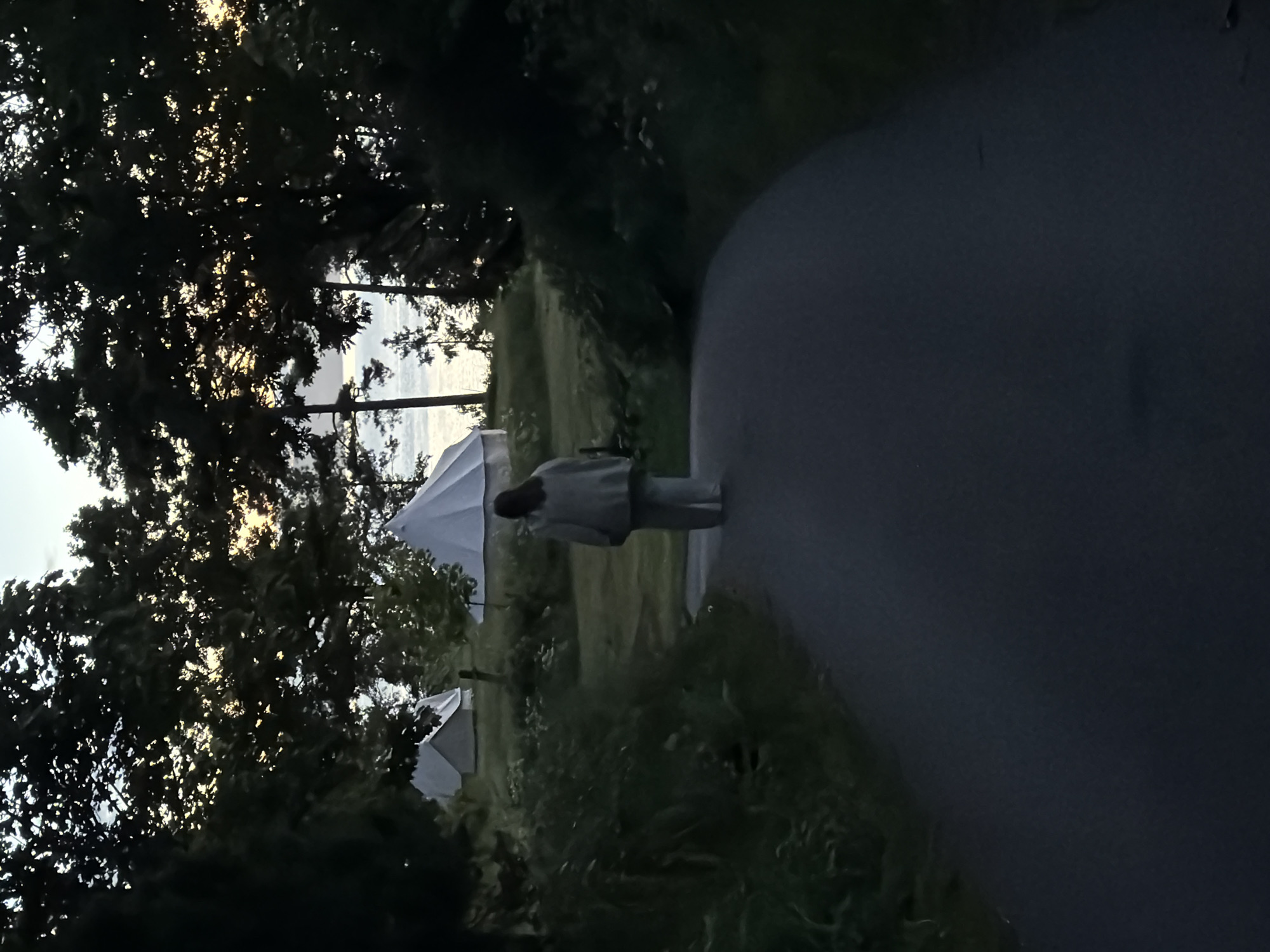 The image captures an outdoor scene at dusk or dawn in Horten, Norway. A single individual, viewed from behind, walks away from the camera on a dark asphalt path that gently curves to the right. The person is dressed in a light-colored, loose-fitting long-sleeved top and matching pants, with long, dark hair falling down their back.
The path is bordered by dark, dense vegetation and bushes. In the middle ground, situated on a grassy area, at least three white, conical-shaped tents are visible, suggesting a glamping or camping environment. Tall, dark-silhouetted trees frame the scene, dominating the upper and side portions of the image. The sky, visible through the tree canopy, is a pale blue-grey with subtle hints of yellow-orange light near the horizon. In the far background, a calm body of water, likely a fjord or sea, is visible between the trees, reflecting the subdued ambient light.