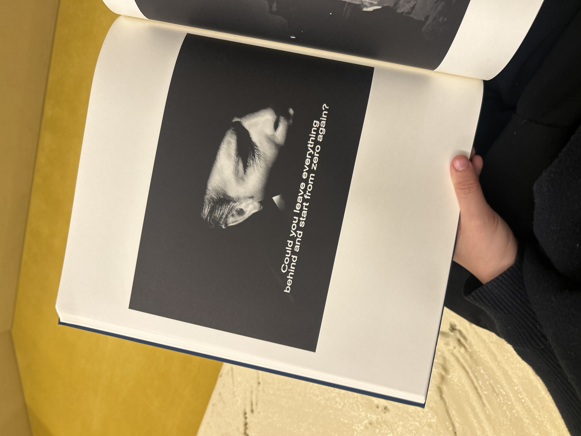 A slightly high-angle shot captures a spread within a book, held open by a hand. The book lies against a backdrop of textured, pale yellow upholstery, possibly a couch or chair. On the left page of the book, a square black-and-white image features a face in profile. Text is overlaid on the photograph, reading "Could you leave everything behind and start from zero again?". The upper page is out of focus.
The image does not contain information that can be used to determine its location in the city of Los Angeles, United States of America.