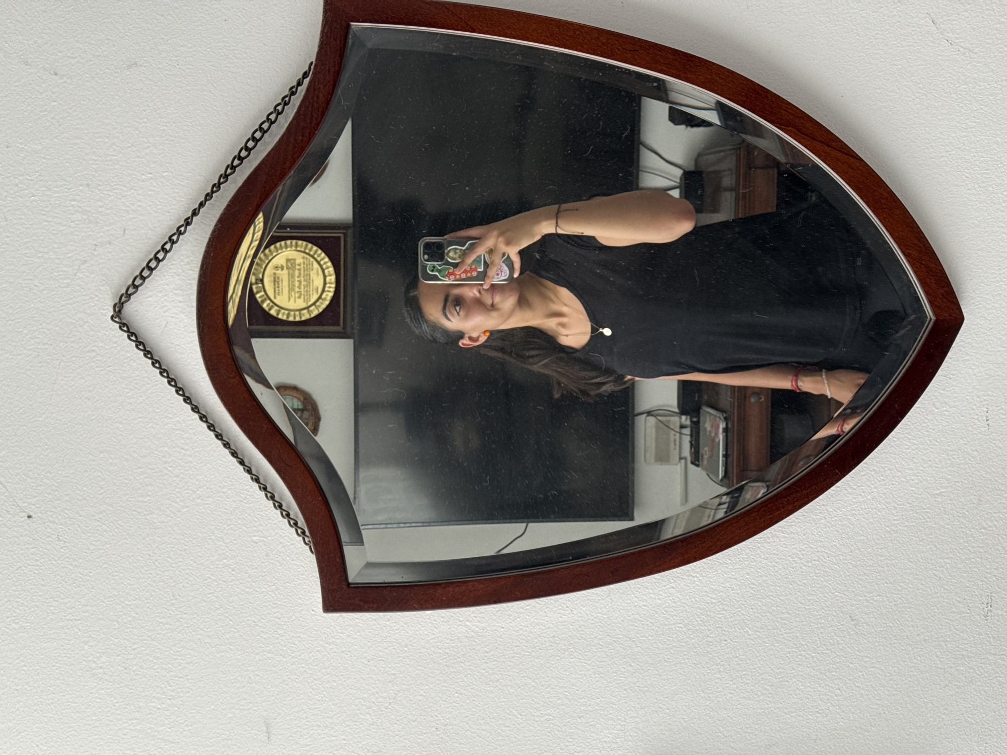 The image shows a woman reflected in an ornate, leaf-shaped mirror. The mirror has a dark wood frame and hangs from a chain against a plain, light-colored wall. The woman is holding a smartphone up to take a selfie. In the background, reflected in the mirror, are furnishings including a television and a cabinet. It is unlikely that the precise location of the image can be determined from the clues in the photograph.
