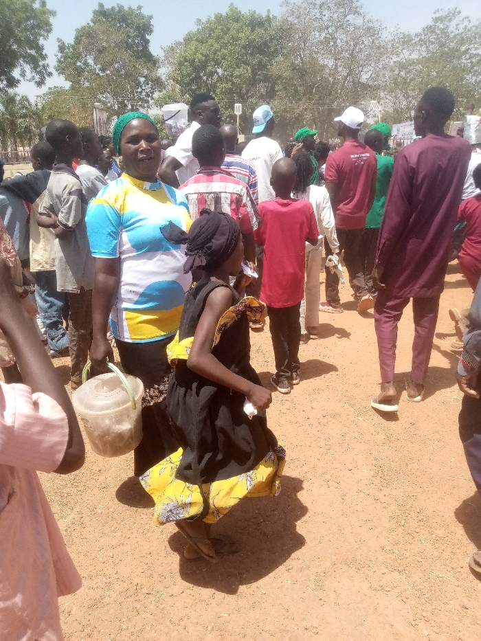 This image depicts an outdoor gathering in Kagoma, Nigeria, occurring on reddish-brown unpaved ground under direct sunlight, indicated by distinct shadows. In the mid-left foreground, a dark-skinned woman wears a green headwrap and a short-sleeved top with blue, white, and yellow patterns. She faces the viewer with a slight smile, holding a transparent plastic bucket containing light-colored material in her left hand. To her right, a dark-skinned child, likely a girl, wears a black sleeveless dress with a wide yellow and blue patterned hem and a black headwrap. The child is captured mid-movement, looking downwards and to the right, holding a small white object in her right hand. Behind these two figures, a dense crowd of individuals, including adults and children, occupies the midground and background. Attire within the crowd varies, with some individuals wearing green shirts/caps, others in red shirts (one displaying "VOLUNTEER" text), white shirts, or patterned clothing. A man in maroon-colored long-sleeved attire stands prominently on the far right, facing away from the viewer. The background features scattered trees with green foliage against a clear, bright sky.
