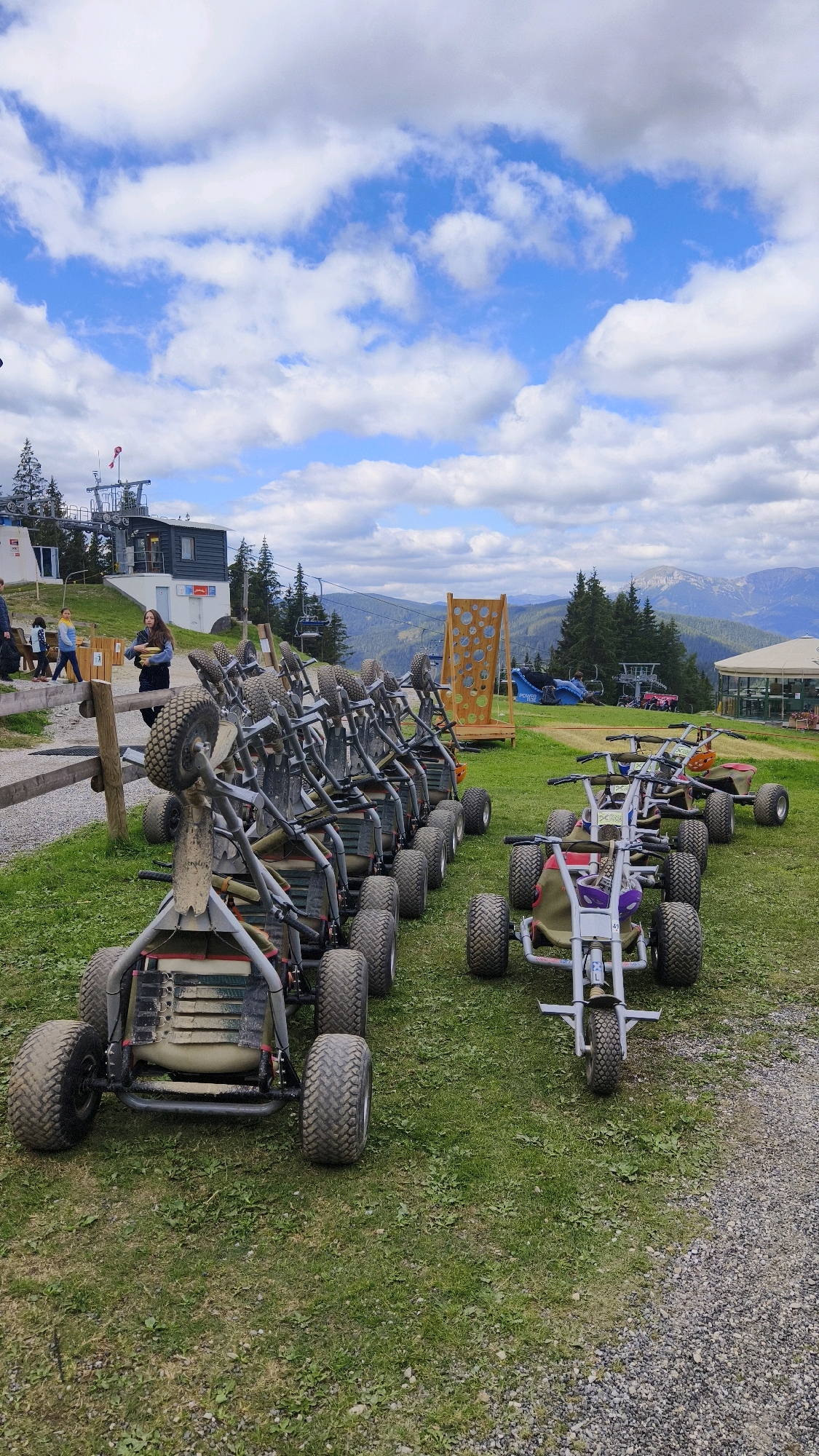 The image captures a view of a mountain landscape with several go-karts parked on a grassy area. In the background, a ski lift structure is visible, suggesting the location is a mountain resort. The sky is partly cloudy with blue patches. To the right of the go-karts, a wooden structure with circular cutouts stands on the grass. Further in the distance, mountains are visible, along with some buildings. There are a few people in the mid-ground, appearing to be walking or standing near the go-karts. The go-karts themselves are small, single-seater vehicles with rugged tires, arranged in rows. The scene appears to be set in an outdoor recreational area in Semmering, Austria, likely a venue for gravity-powered carting or similar activities.
