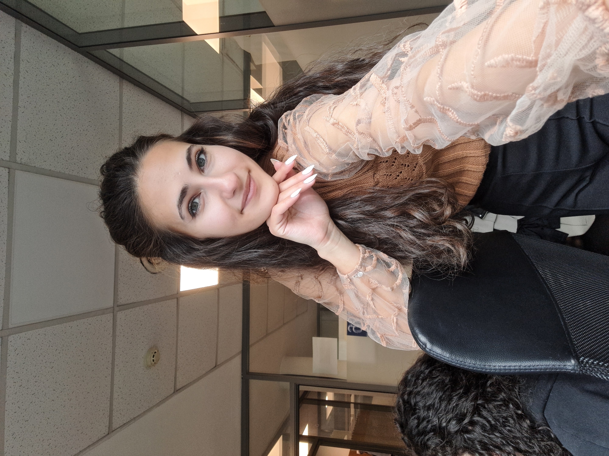 A young woman with long, dark, wavy hair is featured in a portrait-style selfie, looking directly at the camera with a gentle smile. She appears to be in her late teens or early twenties. Her right hand is brought up towards her chin, revealing white, pointed fingernails. She is dressed in a light brown ribbed knit top, layered under a sheer, light peach or nude-colored long-sleeved blouse with elaborate embroidered patterns. Dark pants are visible below her waist.
The setting is an indoor office or commercial building. The background features a ceiling with acoustic tiles, recessed lighting fixtures (one brightly illuminated), and a small white fire alarm or smoke detector. Glass partitions or windows can be seen separating different areas within the building, reflecting some internal structures. A black office chair is partially visible in the lower right corner, with the top of a person's head, featuring dark curly hair, sitting in it. The lighting is predominantly artificial, consistent with an indoor environment. The woman's expression conveys a pleasant and approachable demeanor. This scene is set in Sofia, Bulgaria. No discernible text is visible within the image.