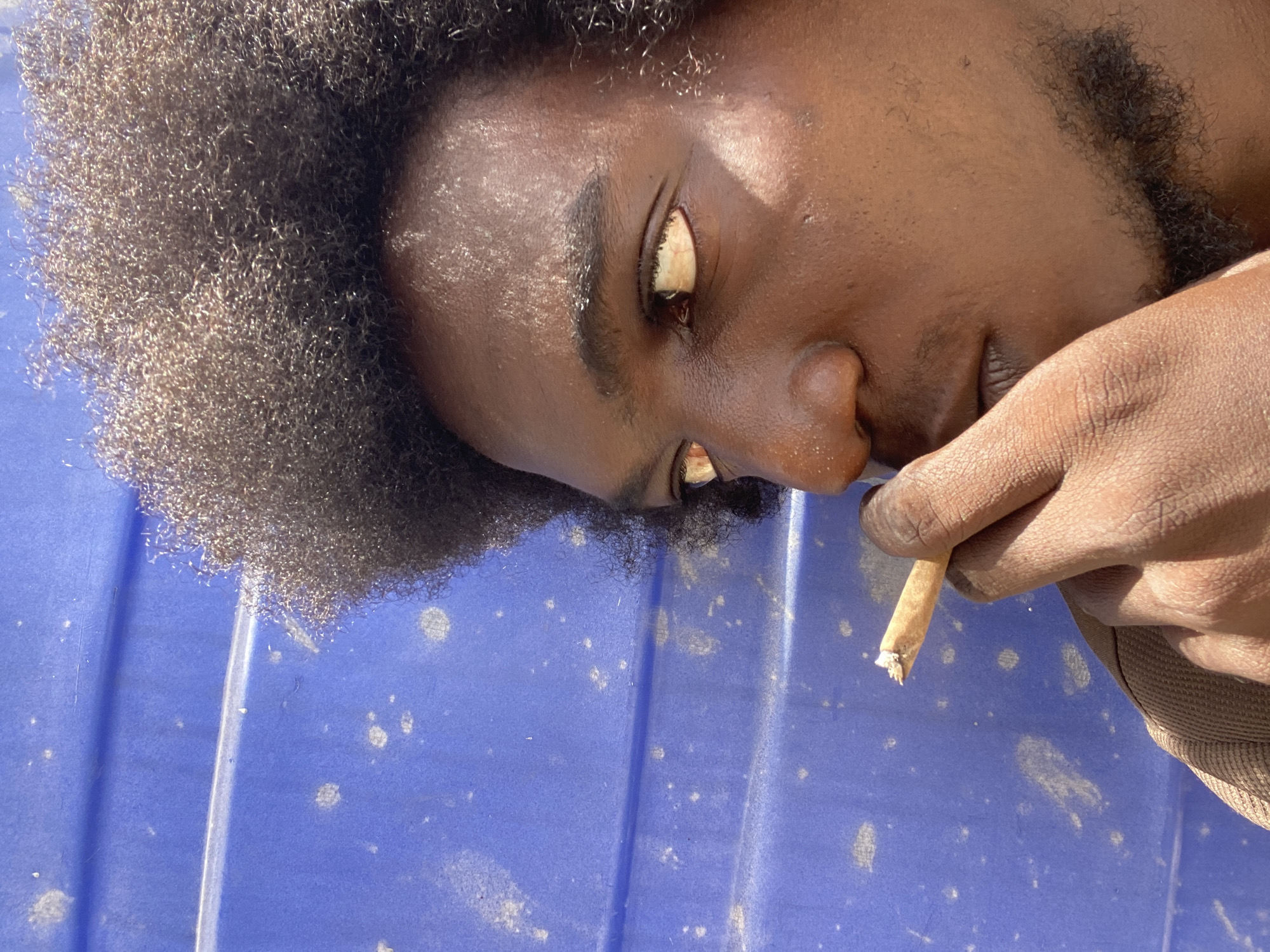 A dark-skinned male with an afro and beard is holding a lit cigarette in his right hand. He is lying on a blue corrugated surface, possibly a bin or similar object. The environment appears to be outdoors, with harsh sunlight suggesting a tropical climate. It is unknown if the subject is in the city of Lagos, Nigeria.
