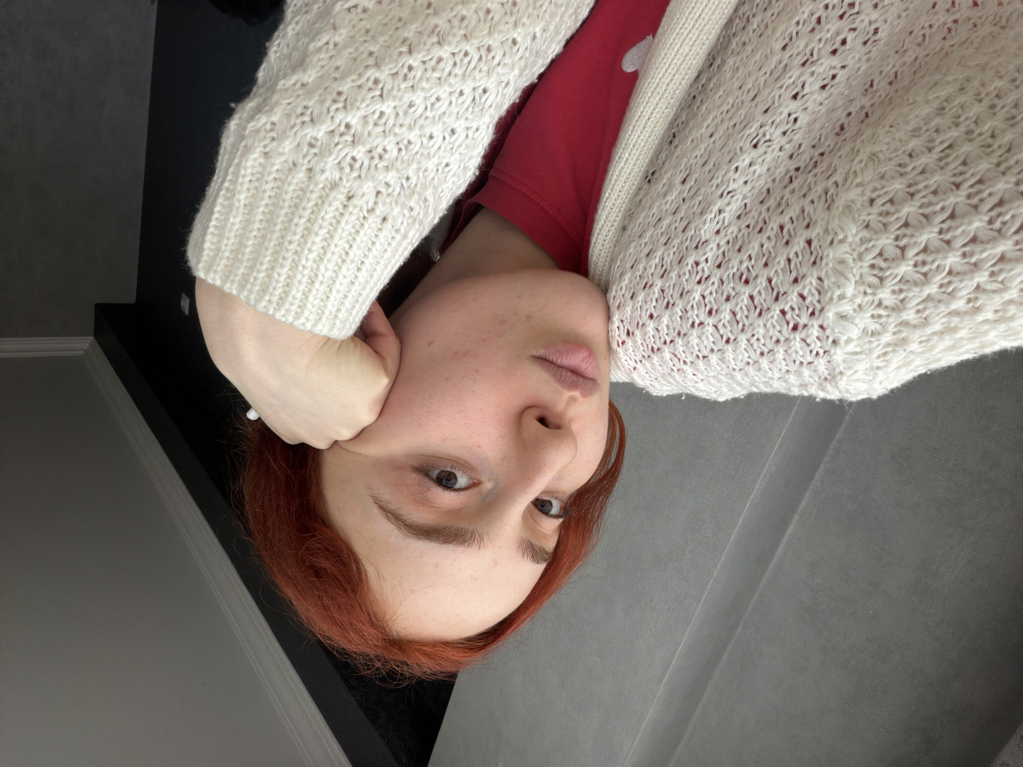 This image, oriented upside down and taken in Tbilisi, Georgia, features a person with reddish-brown hair, likely a woman. She has fair skin with some natural blemishes and dark brown eyes that directly meet the viewer's gaze. Her lips are light pink and slightly parted. She is wearing a plain red top beneath a cream-colored, open-knit cardigan. The cardigan displays a distinct, intricate lace-like or openwork pattern on its body and a ribbed cuff on the sleeve. Her right hand, forming a partly closed fist, supports the side of her face, suggesting a relaxed or pensive pose.
The background indicates an indoor setting, composed of dark, geometric surfaces. A dark gray or black vertical panel is visible on the left, while the right side shows a lighter gray or black surface with a clear diagonal seam or edge, possibly indicating a wall junction or furniture piece. The lighting appears soft and indirect. There is no visible text.
