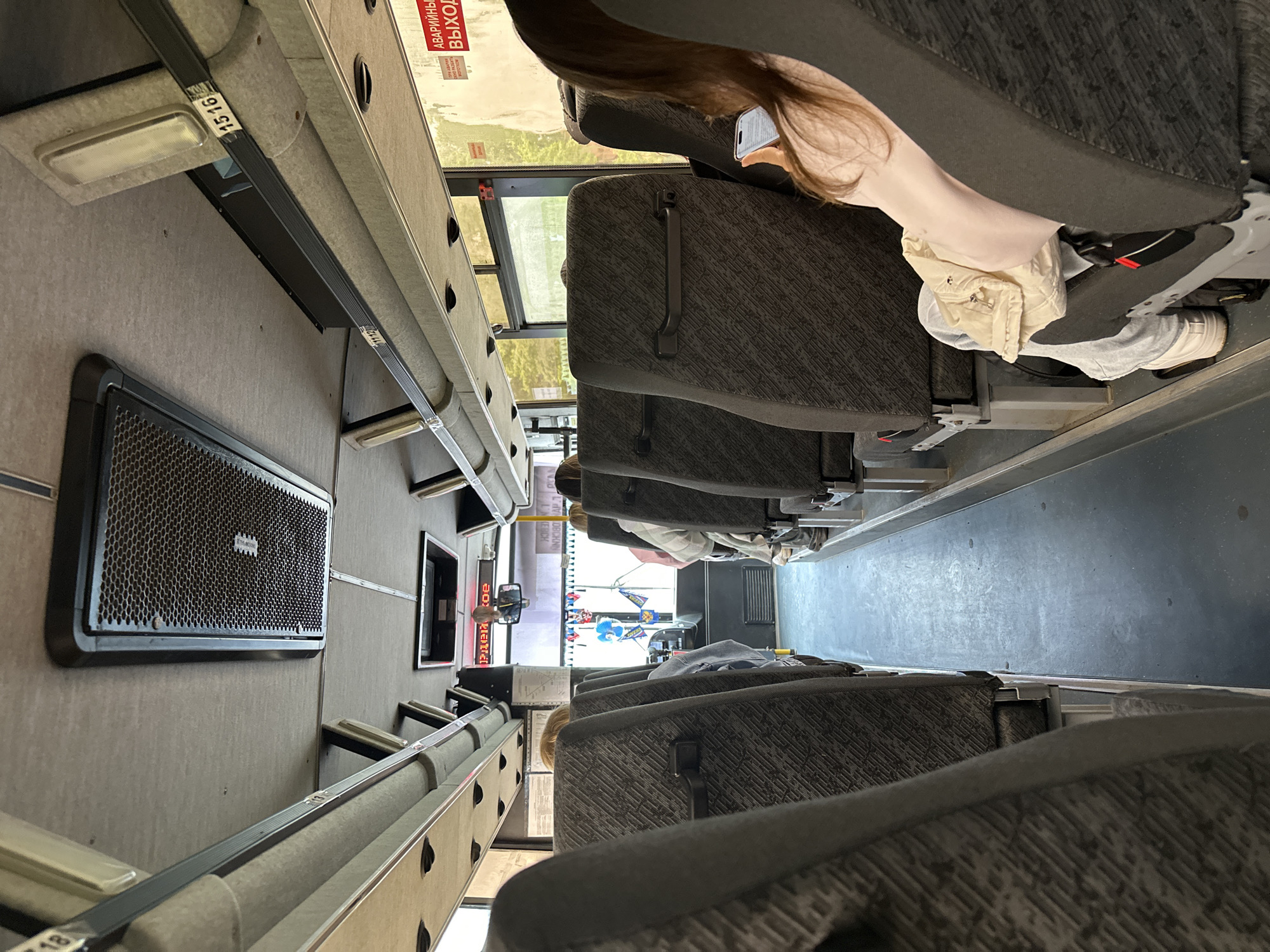 This image captures the interior of a passenger bus or coach, presumably in Chaikovsky, Russia. The perspective is from a seat further back, looking forward down the central aisle towards the driver's area.
The bus interior features rows of high-backed seats upholstered in a dark gray fabric with a subtle geometric pattern. Dark gray plastic handles are affixed to the back of each seat. An aisle with a dark gray floor runs between the two rows of seats. Overhead, the ceiling is covered in a light gray fabric, featuring rectangular ventilation grilles, some with a visible metallic mesh, and elongated rectangular light fixtures. Overhead luggage racks, also covered in the light gray fabric, display small numerical labels such as "11 12", "15 16", and "18".
Several passengers are visible. In the immediate foreground on the right, a person with long brown hair, wearing a light pink top, is seated and engaged with a white smartphone held in both hands. A light-colored jacket is draped over their lap, and they wear light gray trousers and white sneakers. Further forward on both sides of the aisle, other passengers are seated, their heads partially obscured by the seatbacks. One passenger on the left appears to have light-colored hair.
On the right side of the bus, a large window reveals blurred green foliage and possibly some sunlight outside. A prominent red sign on the window or wall reads "АВАРИЙНЫЙ ВЫХОД" (EMERGENCY EXIT), with smaller white text above it stating "ПРИ АВАРИИ РАЗБИТЬ МОЛОТКОМ" (IN CASE OF EMERGENCY BREAK WITH HAMMER).
At the front of the bus, the driver's area is partially visible. An electronic display above the windshield shows red, inverted text, possibly reading "РАСПОРЯДОК" (ROUTINE/SCHEDULE) or a similar word. A rearview mirror is mounted centrally. Near the driver's station, small decorative flags in blue, yellow, and white, along with a blue balloon-like object, are visible. On the left side near the front, a white information board with diagrams and text is mounted.