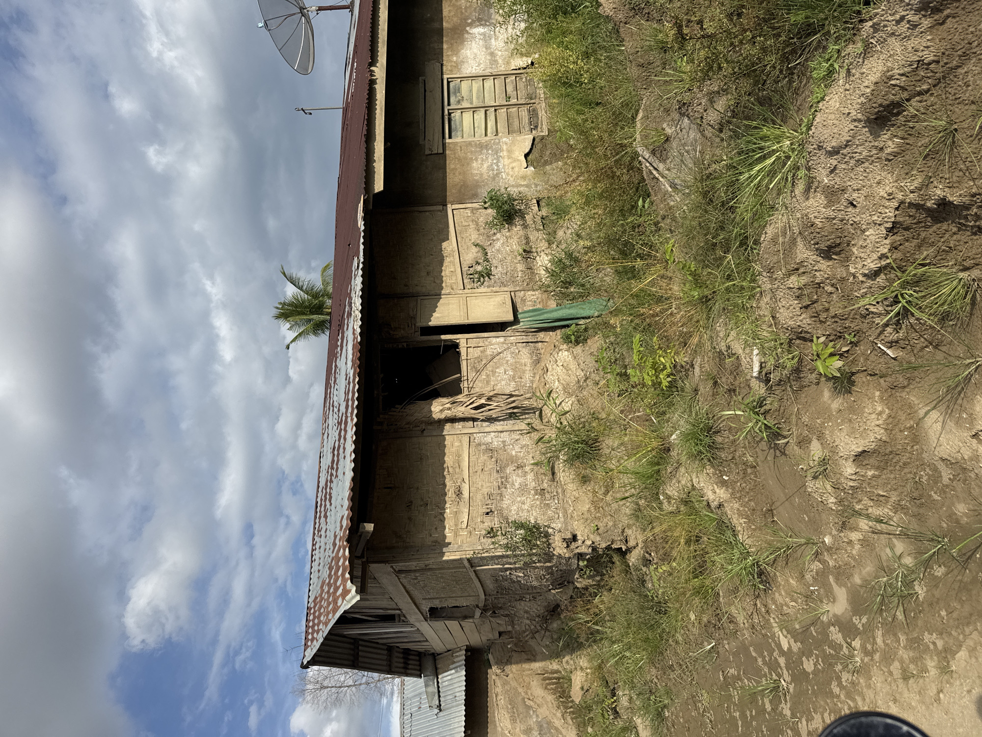 The image shows a dilapidated building with a corrugated metal roof, likely in Gampong Blang, Indonesia. The walls appear to be constructed from a combination of mud, wood, and woven material, with sections showing significant damage and erosion. One window is open with wooden shutters, and another has a louvered window with broken panes. Overgrown vegetation surrounds the structure, with patches of bare earth and mud in the foreground. The sky is partly cloudy. A satellite dish is visible on the roof of an adjacent building.