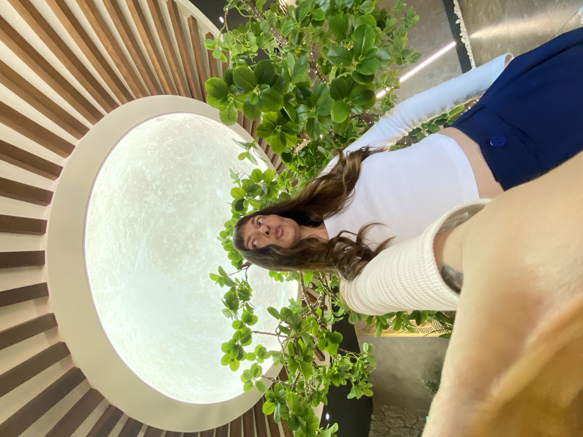 An indoor shot captures a woman taking a selfie from a low angle, looking upwards and to the right. She has long, dark hair and is wearing a white long-sleeved shirt and dark blue pants. She is making a playful kissing face. Behind her, there is a large, circular light fixture resembling the moon, with wooden slats radiating outwards from it, suggesting a modern ceiling design. Artificial green foliage is visible, framing the woman and the moon-like light. The background appears to be an interior space with a stone-like wall texture and illuminated strips on the floor. The scene is brightly lit, focusing on the woman and the overhead feature. The image does not provide information about the city of Votkinsk, Russia.