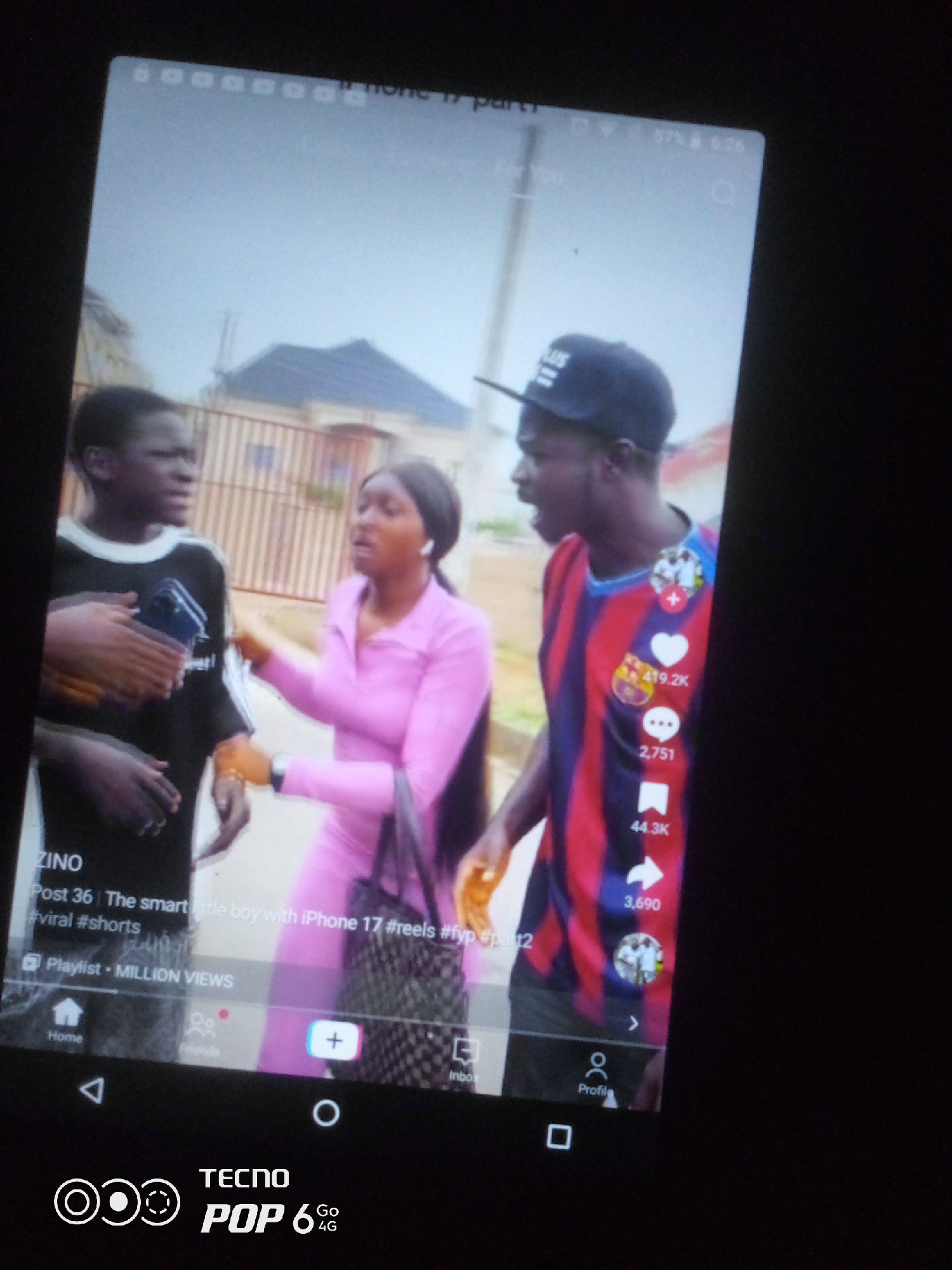 A mobile phone screen displays a video of three young people in an outdoor urban setting, possibly in the city of Ilorin, Nigeria, given the presence of the TECNO POP 6 4G branding, a popular mobile phone brand in Nigeria. The video appears to be a TikTok or similar short-form content. In the foreground, a person with dark skin, wearing a black t-shirt with white stripes on the sleeves, is holding a blue object, possibly a phone, and looking to their right. To their right, a young woman in a bright pink long-sleeved outfit and matching pants is gesturing with her right hand towards the person with the black t-shirt, her mouth open as if speaking. Her left arm is bent, and she is holding a patterned bag. Further to the right, a young man wearing a black baseball cap and a horizontally striped t-shirt in red, blue, and black is looking towards the woman and speaking, with his mouth open. Social media engagement metrics such as likes (419.2K), comments (2,751), and shares (44.3K) are visible on the right side of the screen. The background shows a building with a tiled roof and a fence, suggesting a residential area. The bottom of the phone screen displays navigation icons for "Home," "Friends," a plus sign for creating content, and "Inbox," along with the TECNO POP 6 4G logo. The overall scene captured in the video is dynamic, with animated gestures and expressions suggesting a conversation or a playful interaction.