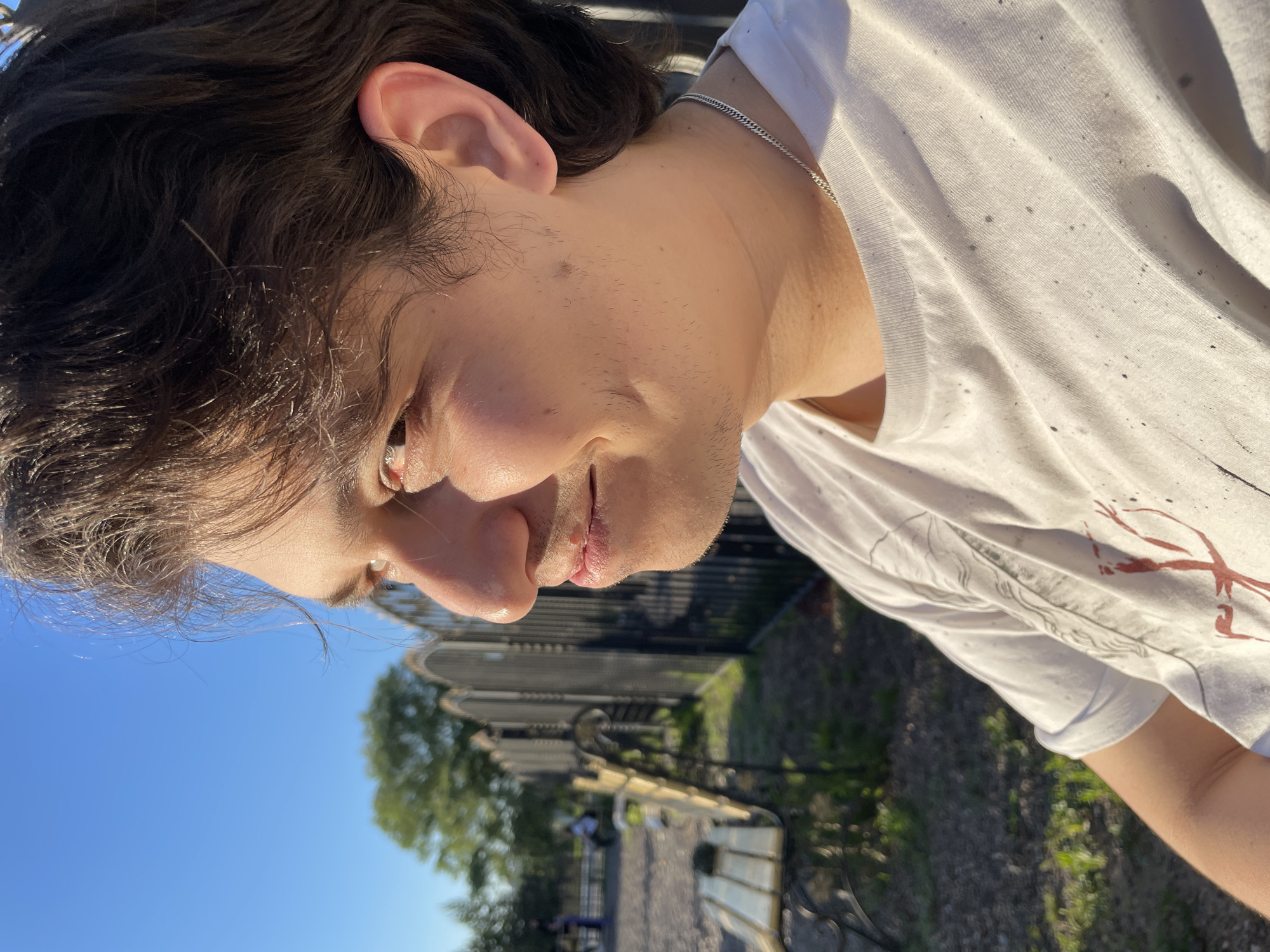A close-up, profile shot of a young man sitting outdoors in what appears to be a park. The man has dark, wavy hair and is wearing a white t-shirt with a silver chain. He is looking off to the left, with a slight smile on his face. Sunlight is illuminating his face, creating highlights. In the background, a black metal fence separates the park area from a path and a bench. There are trees and a clear blue sky. The overall impression is of a casual, sunny day in a public space. Without further information or context, it is impossible to determine the specific location within Izhevsk, Russia.