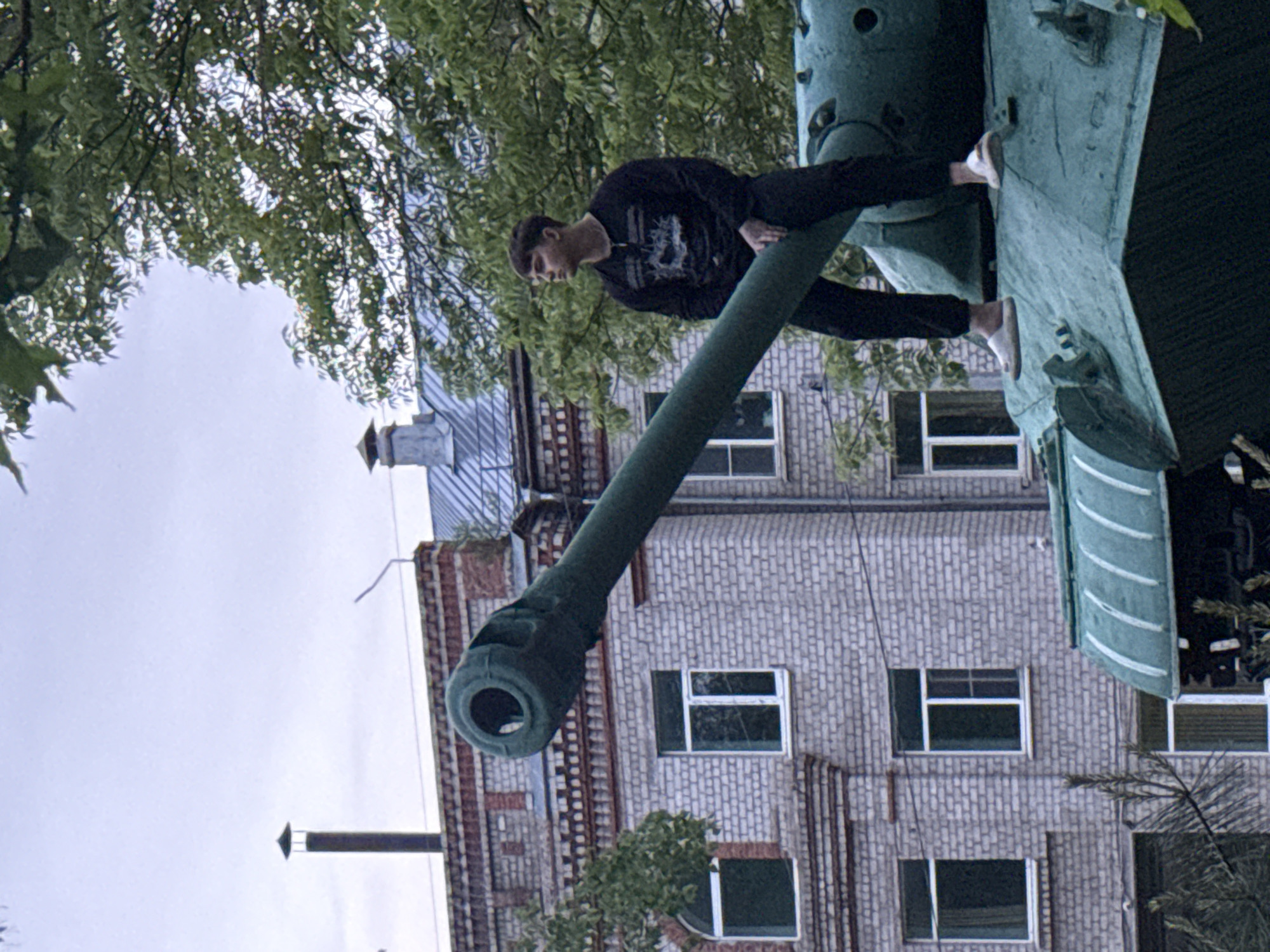 An outdoor photograph, likely taken in Birobidzhan, Russia, captures a young man posing on a military tank, which appears to be a monument. The tank, painted in a dark teal or green, features a long main gun barrel and a visible turret. The young man, dressed in a black long-sleeved shirt with a light-colored graphic design and dark pants, is balancing on the gun barrel and a section of the tank's turret. He wears light-colored footwear, possibly white shoes or slippers, and is looking slightly downwards.
In the background, a multi-story brick building with a reddish-brown hue and numerous rectangular windows is prominent. The building exhibits some architectural detailing, including decorative brickwork. The sky is overcast and light, indicating daytime without direct sunlight. Lush green tree foliage partially frames the scene, obscuring parts of the sky and the building, contributing to an urban or suburban setting with natural elements. The overall atmosphere suggests a casual moment captured at a public display or historical site. No discernible text is visible on any of the subjects or objects in the image.