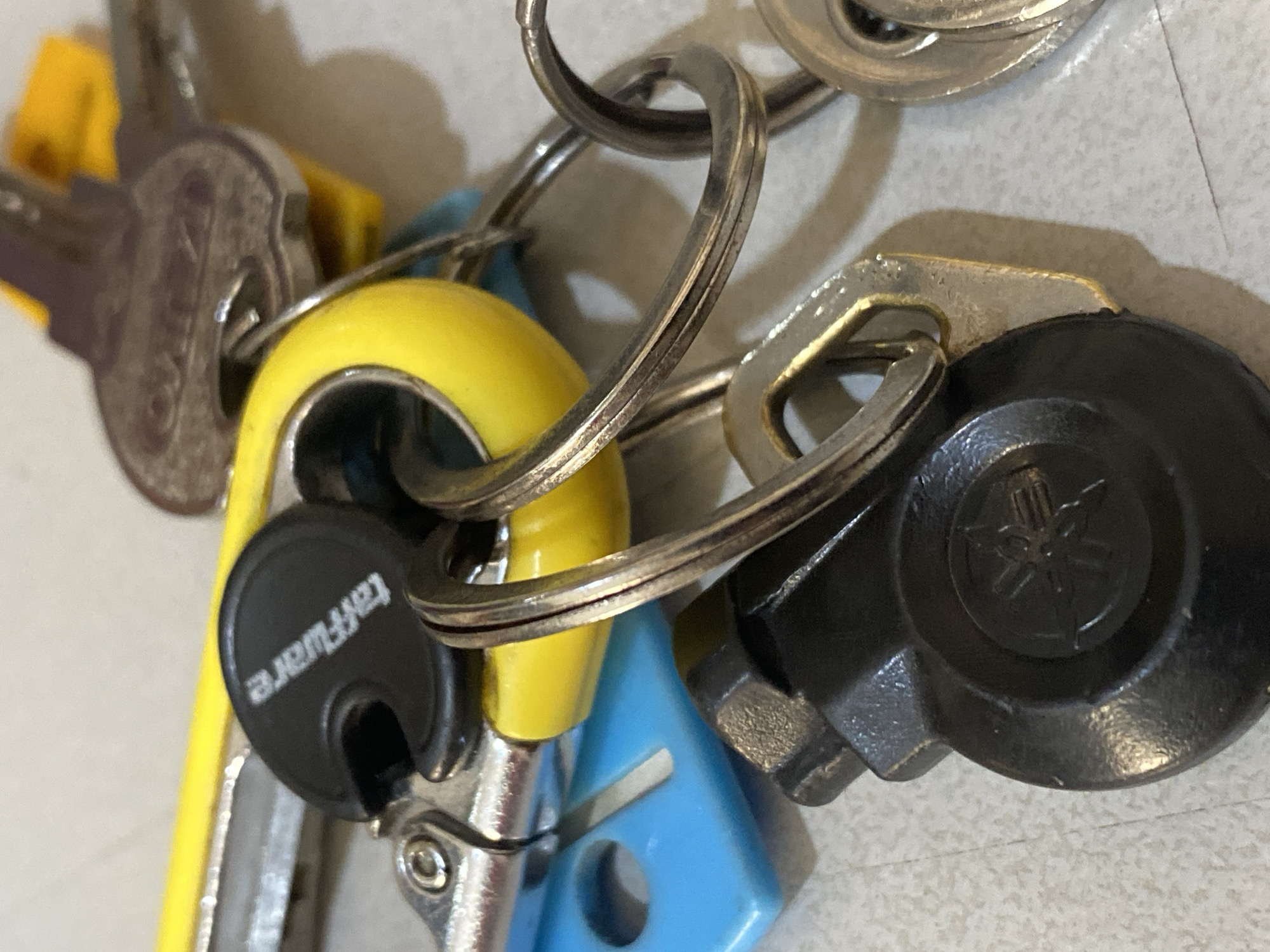 A close-up, high-angle shot shows a keychain with multiple keys, a blue plastic tag, a yellow object, and a black disc with a Yamaha logo. The keychain is on a light-colored, speckled surface, possibly a tiled floor. The image does not provide any information about the city of North Jakarta, Indonesia, or any individuals present.