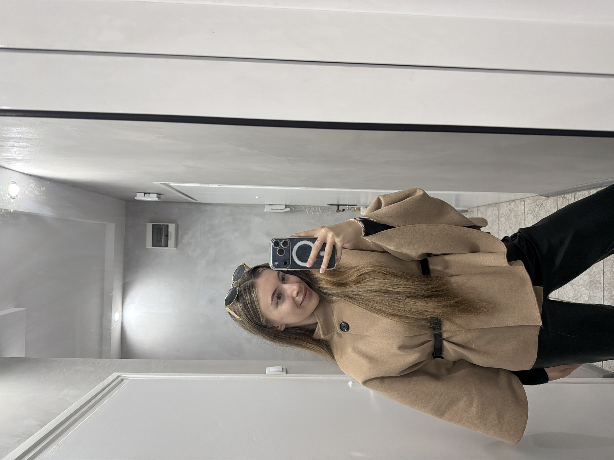 A woman is taking a selfie in what appears to be an interior hallway or possibly a foyer of a building. The walls are painted in a light grey, and the trim and doors are painted white. The woman is wearing a tan coat, black pants, and sunglasses. The coat is belted at the waist. She is holding a cell phone in her right hand to capture the photograph. The background is somewhat blurred, but there is a light fixture visible on the wall behind her. The location is unknown, but it is specified it should be in the city of Plovdiv, Bulgaria. Since there are no specific features to determine the location, this cannot be confirmed.