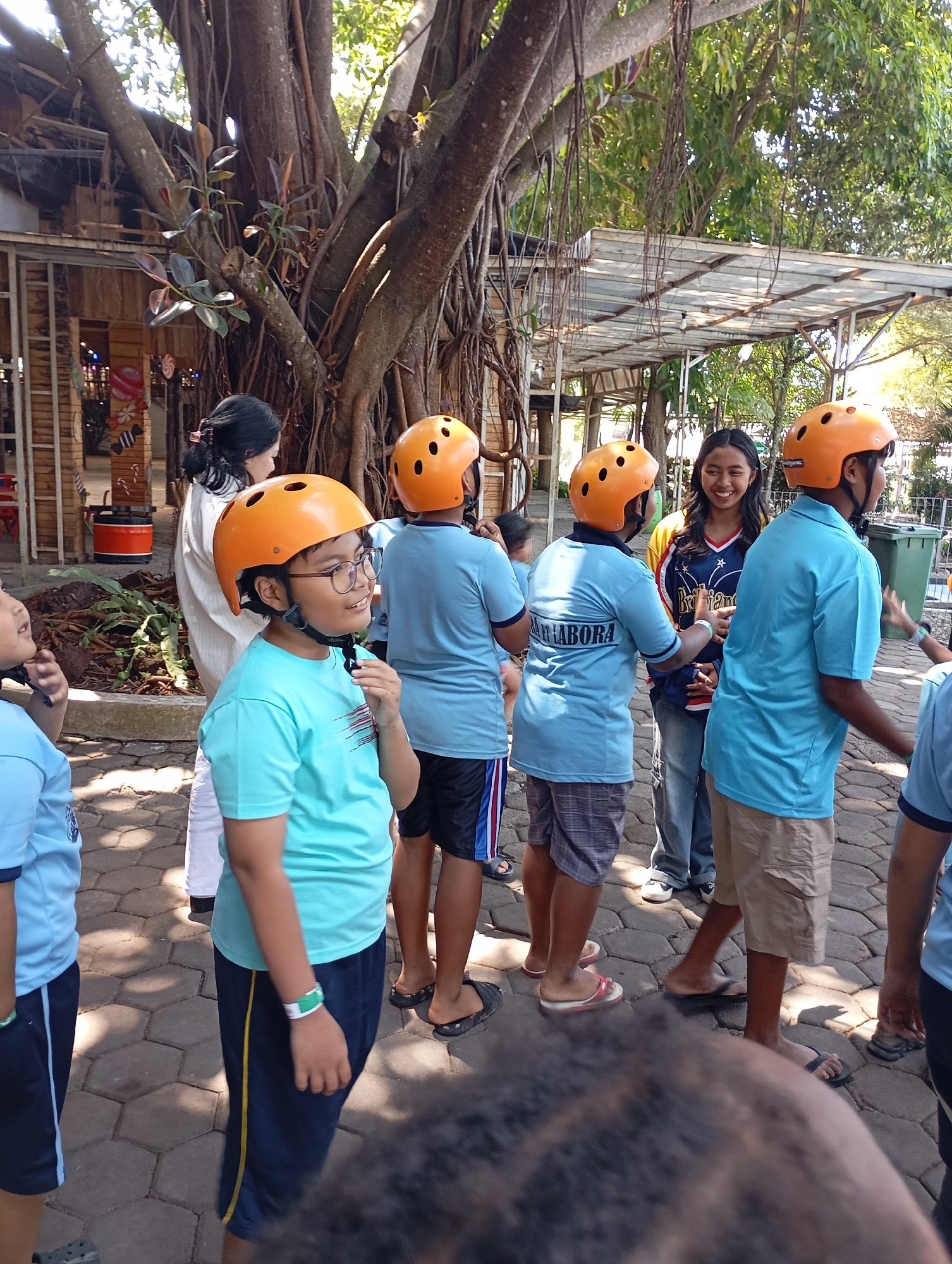 In Batu, Indonesia, a group of people, primarily children, are gathered outdoors, near a large banyan tree. Most of the children are wearing bright orange helmets and light blue shirts, suggesting they are participating in an activity. One child in the foreground, wearing a blue shirt and an orange helmet, is looking to the side and touching their chin. Another child in a blue shirt and orange helmet is also visible. A young woman in the background is smiling and looking at the children. The environment appears to be a park or recreational area, with paved ground and some greenery. The overall scene suggests a fun and engaging outdoor event for children.