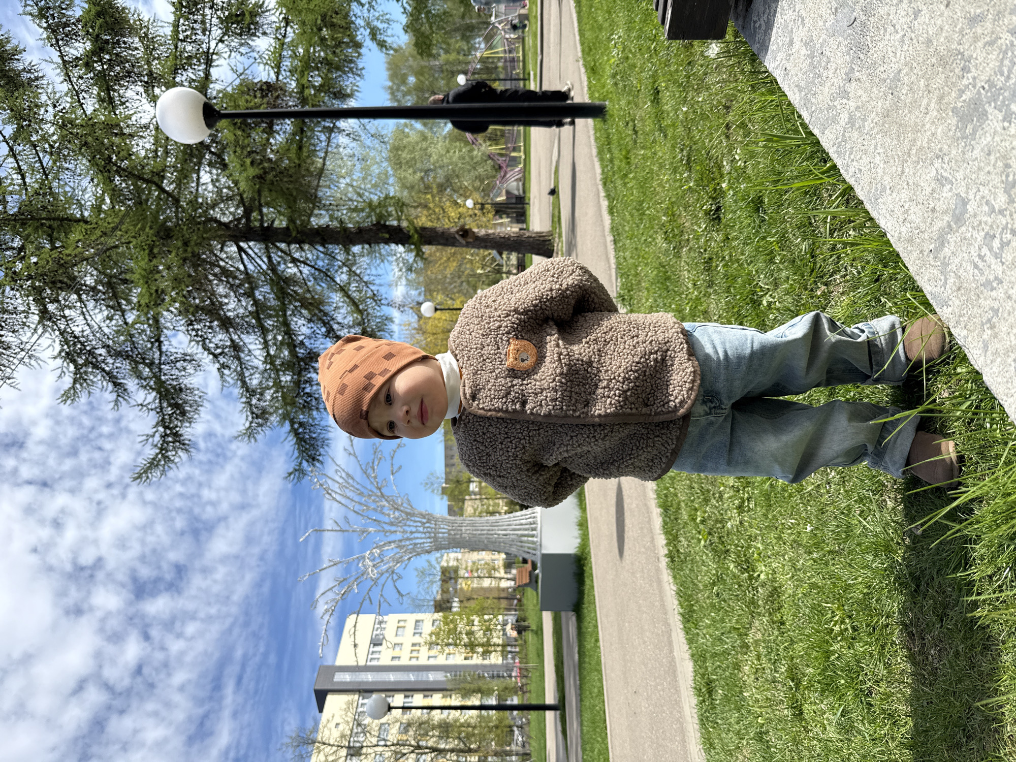 A child stands in a park on a sunny day in Unknown, Unknown. The child wears a brown hat, a brown jacket, and blue jeans. They are standing on green grass next to a paved path and a low concrete wall. In the background, there are trees, street lights, buildings, and a white decorative tree structure. A person walks in the distance. The sky is blue with scattered clouds.