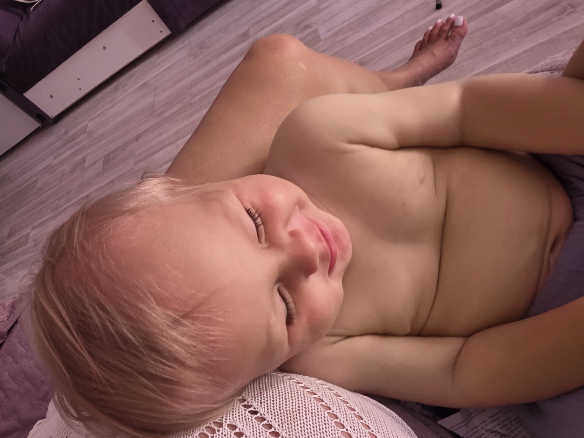 The image shows a close-up, high-angle shot of a baby lying on their side. The baby has fair hair and is partially undressed, with their chest and abdomen visible. Their eyes are closed, and they have a content expression, with rosy cheeks. The baby is resting on a textured, light-colored blanket or textile. In the background, a person's bare leg and foot are visible, with white-painted toenails. The person's leg is positioned behind and to the side of the baby. The floor beneath appears to be light-colored wood-plank flooring, with a slight pinkish hue. A dark purple object, possibly a bed or sofa, is partially visible in the upper left corner of the image. The overall lighting suggests an indoor setting, possibly during the daytime. No visible text is present in the image. The setting is implied to be Sochi, Russia.