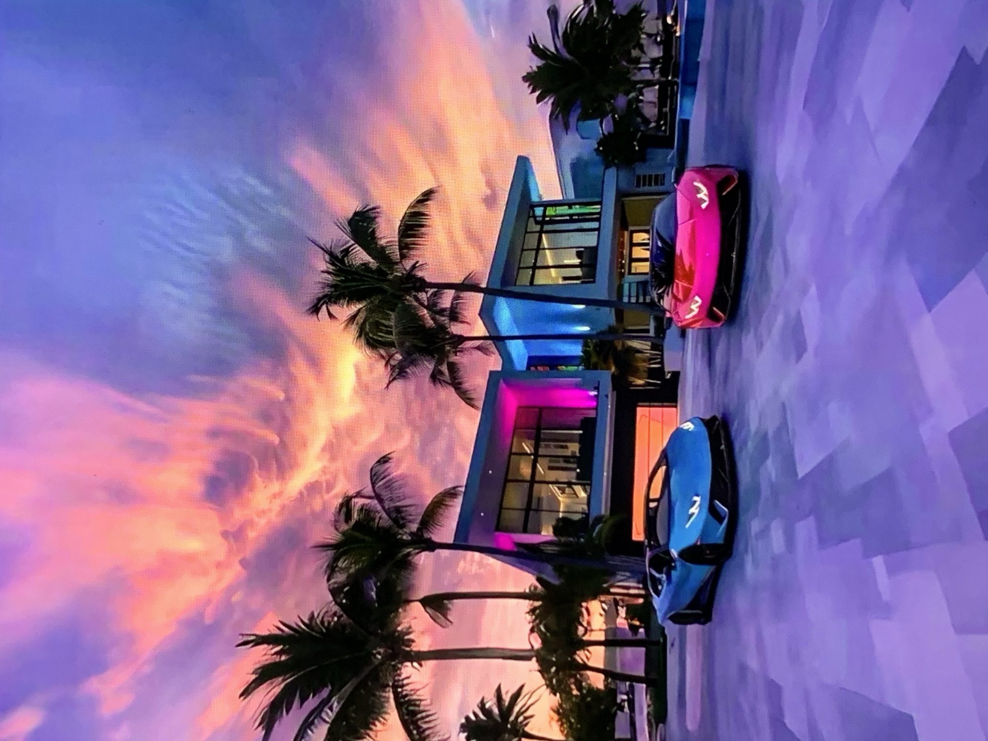 An aerial shot of two luxury sports cars, a blue Lamborghini and a pink Lamborghini, parked on a driveway in front of a modern, multi-story house. The house features large glass windows and is illuminated with vibrant pink and blue lights. Several palm trees are scattered around the property, and the sky above is a dramatic sunset with streaks of orange, pink, and purple clouds. The ground in front of the house is paved with large, irregularly shaped tiles in shades of purple and grey.