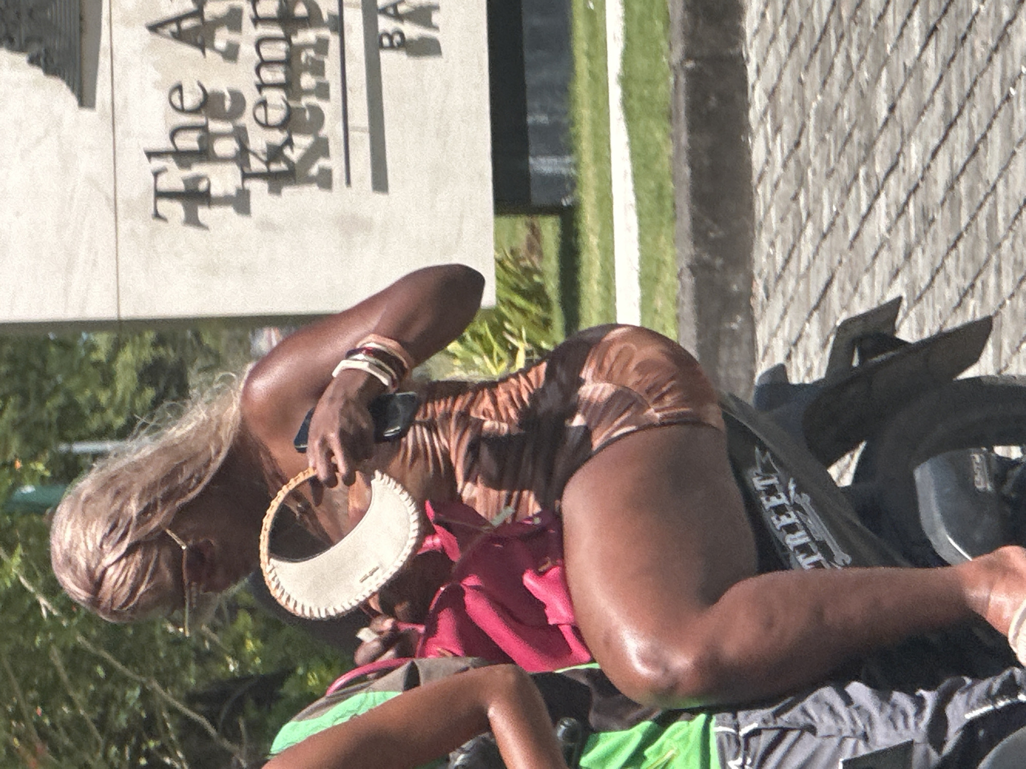 The image shows a person with fair hair and a dark complexion lying on a motorcycle on a sidewalk. The person wears a brown patterned outfit, a sun visor, and gold-rimmed glasses. A red bag is visible between the person and the motorcycle. A sign reading "The Kempinski Bali" is in the background, indicating the location is likely in Kuta Selatan, Indonesia. The environment includes greenery, paving stones, and a glimpse of a green lawn behind a fence.