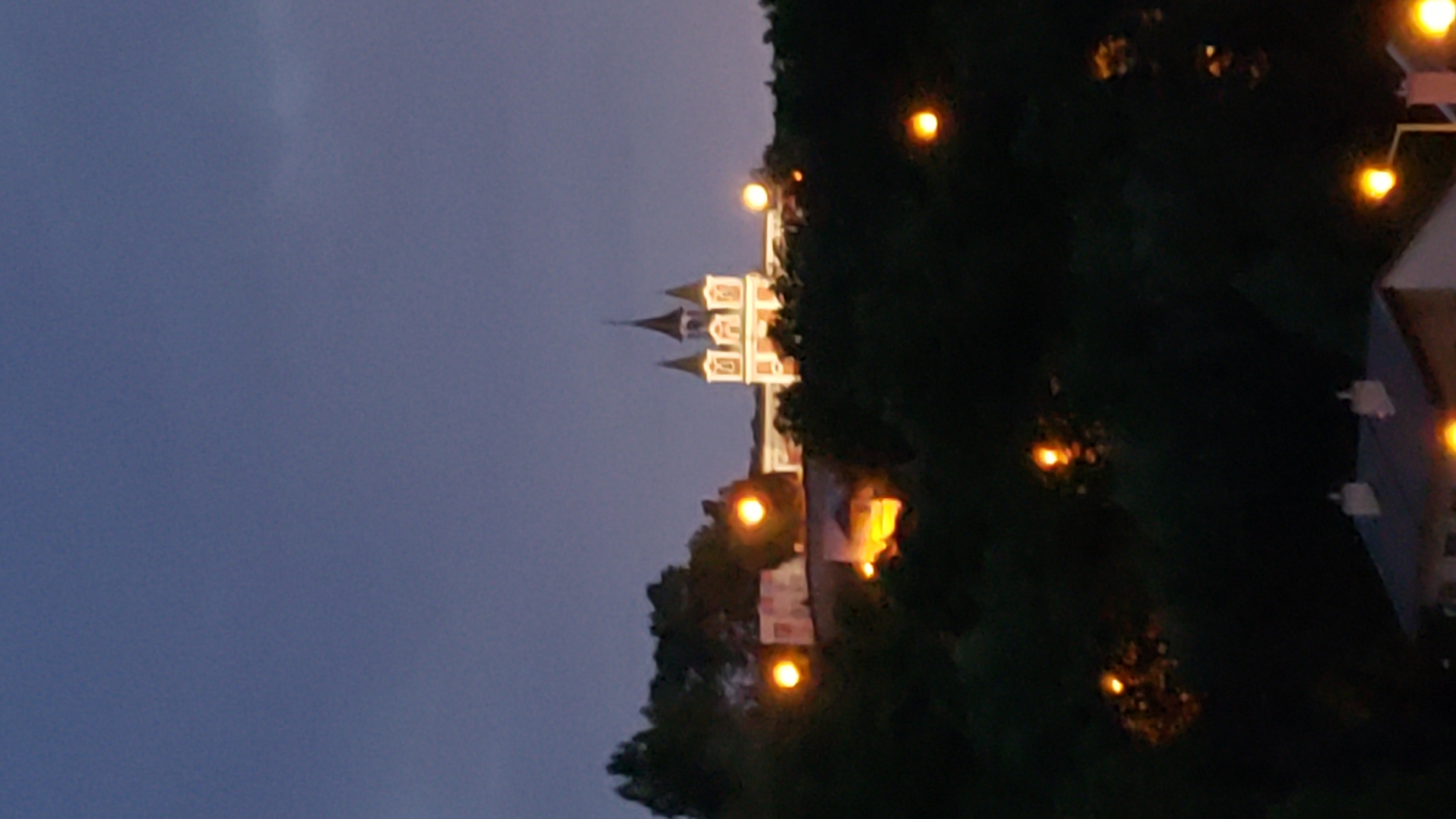 The image depicts a view of the St. Nicholas Cathedral in Prešov, Slovakia, at dusk. The cathedral, a prominent landmark, is illuminated and appears in the upper center of the frame. It features multiple spires, characteristic of its architectural style. Surrounding the cathedral and in the foreground are dark silhouettes of trees. Scattered throughout the image are glowing orbs of light, likely streetlights or house lights, which create a bokeh effect against the dark foliage and twilight sky. A portion of a building with a roof and chimneys is visible in the bottom of the frame, also lit by artificial lights. The sky is a deep blue, transitioning from a lighter hue at the horizon to a darker shade at the top, indicating the late hour. No people are visible in the scene.