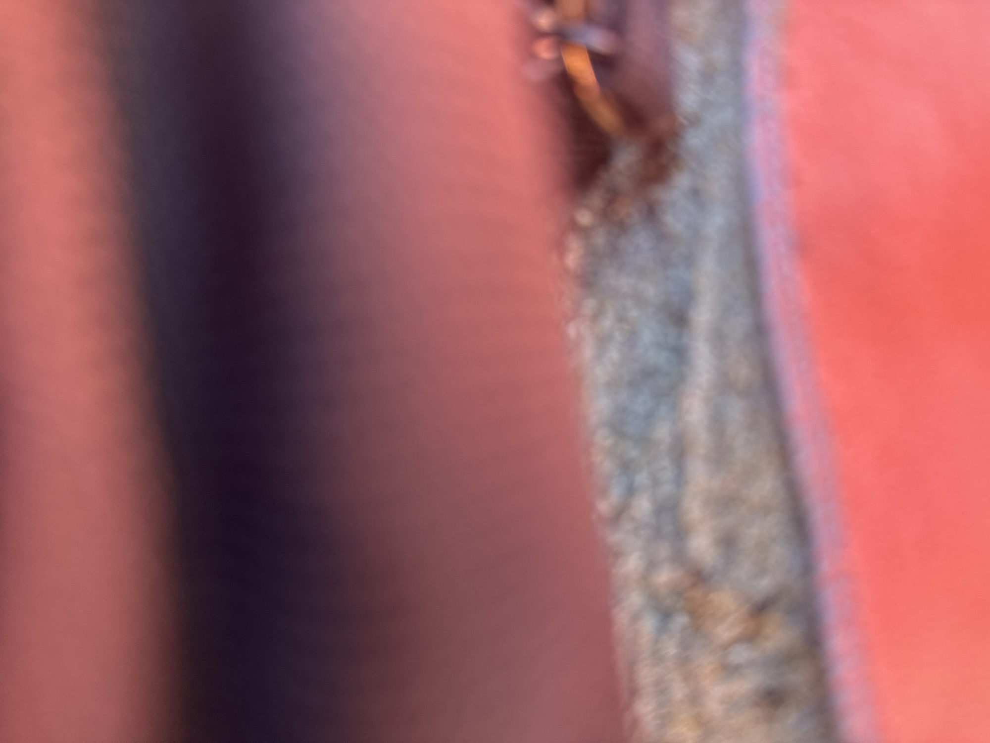 The image shows a very close-up, out-of-focus view of a textured surface, possibly fabric or a rough material, in a reddish-pink color. A golden ring with a slight orange hue is visible on this surface, positioned towards the right side of the frame. Above this textured area, the image transitions to a blurred expanse of darker, muted colors, with shades of purple and darker pink dominating. The overall impression is of an abstract or detail-oriented shot, lacking clear contextual elements. There are no discernible people, actions, or environmental features that would allow for localization within Kranevo, Bulgaria, or any other specific geographic area.