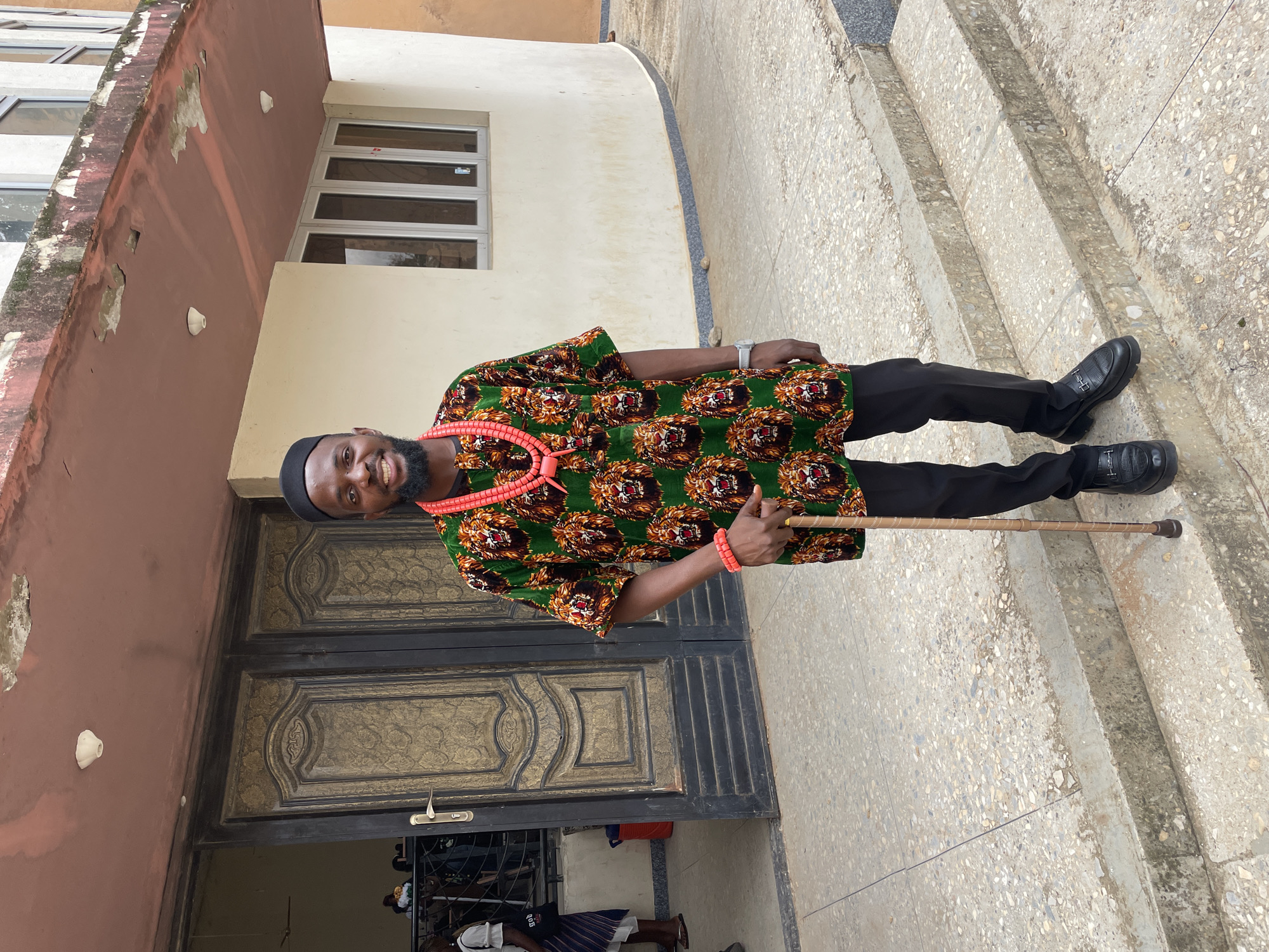 A man stands on a concrete staircase outside a building with large double doors. He is wearing a green and black patterned shirt with lion faces, black trousers, black shoes, and a black cap. He is also wearing red beaded necklaces and a red beaded bracelet on his left wrist. He is holding a walking stick with a brown handle. The building appears to be made of concrete and has a red trim. The doors are dark brown with intricate gold detailing. The background shows other people and architectural elements. The scene appears to be in Jos, Nigeria.