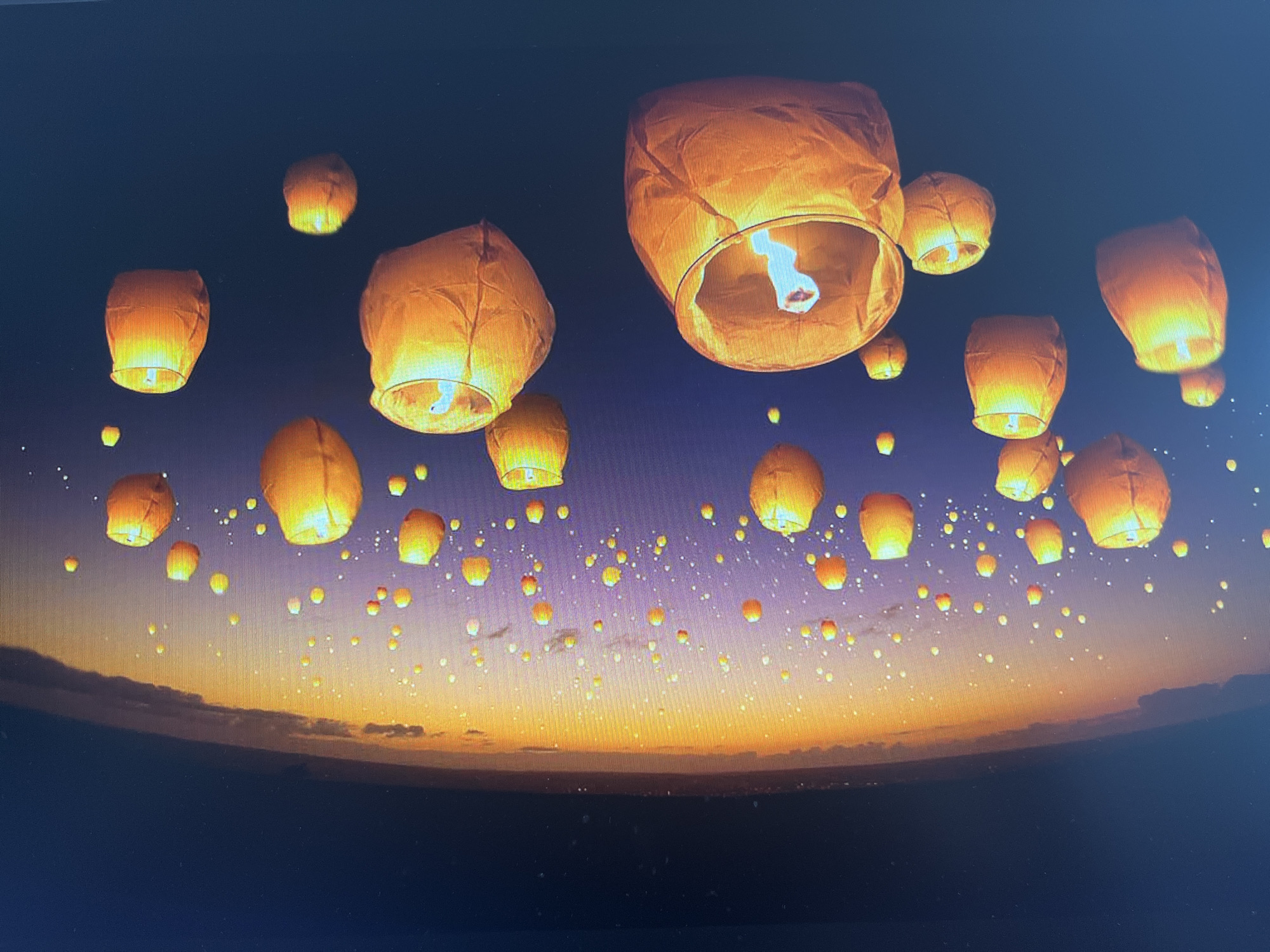 The image displays a panoramic view of a night sky above Varna, Bulgaria, illuminated by countless sky lanterns. The primary subjects are hundreds of glowing, orange and yellow paper lanterns of varying sizes, floating upwards. Many larger lanterns are visible in the foreground and mid-ground, clearly showing the internal flame that provides their illumination.
The scene depicts dusk or early night, with the upper sky a deep dark blue to black, gradually transitioning into a vibrant palette of orange, yellow, and purple along the horizon. This suggests the last light of a sunset. Below the horizon, the landscape or sea is dark and indistinct. The smaller, more distant lanterns appear as numerous bright specks, creating a dense constellation-like pattern across the sky, extending down to the colorful horizon.
The activity taking place is a mass release of these lanterns, indicating a festival, celebration, or a significant event. The clear sky allows for unobstructed visibility of the lanterns and the dramatic colors of the sunset. There is no visible text in the image. The overall impression is one of magic, beauty, and solemn celebration.