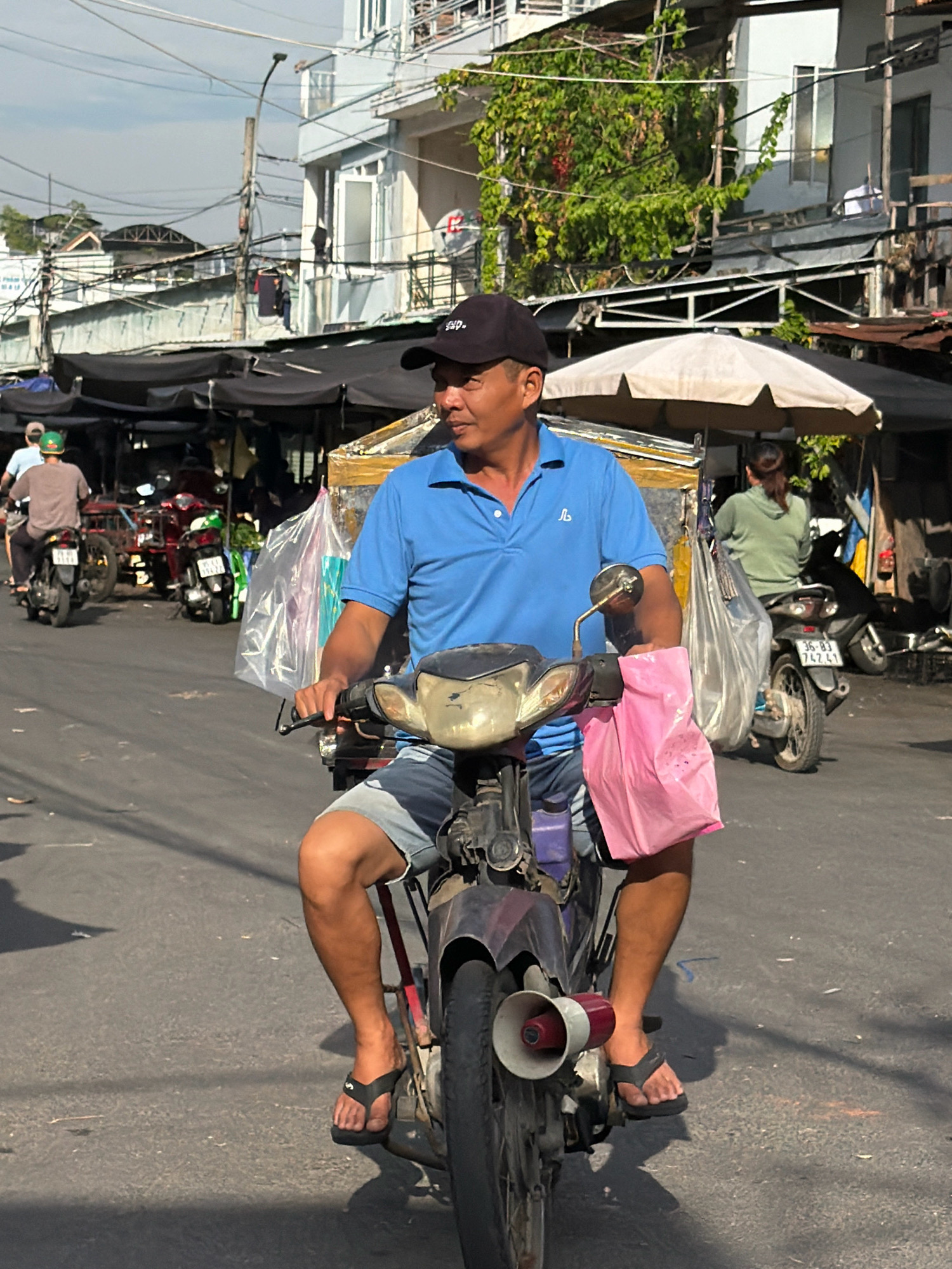 In Nha Trang, Vietnam, a man is riding a motorcycle down a street. He is wearing a blue polo shirt, denim shorts, black flip-flops, and a black baseball cap. He is carrying a pink plastic bag on the back of his motorcycle, and a clear plastic bag is also attached. The man is looking to his left. The street is lined with market stalls and buildings. There are other motorcycles and people in the background. The sky is cloudy.