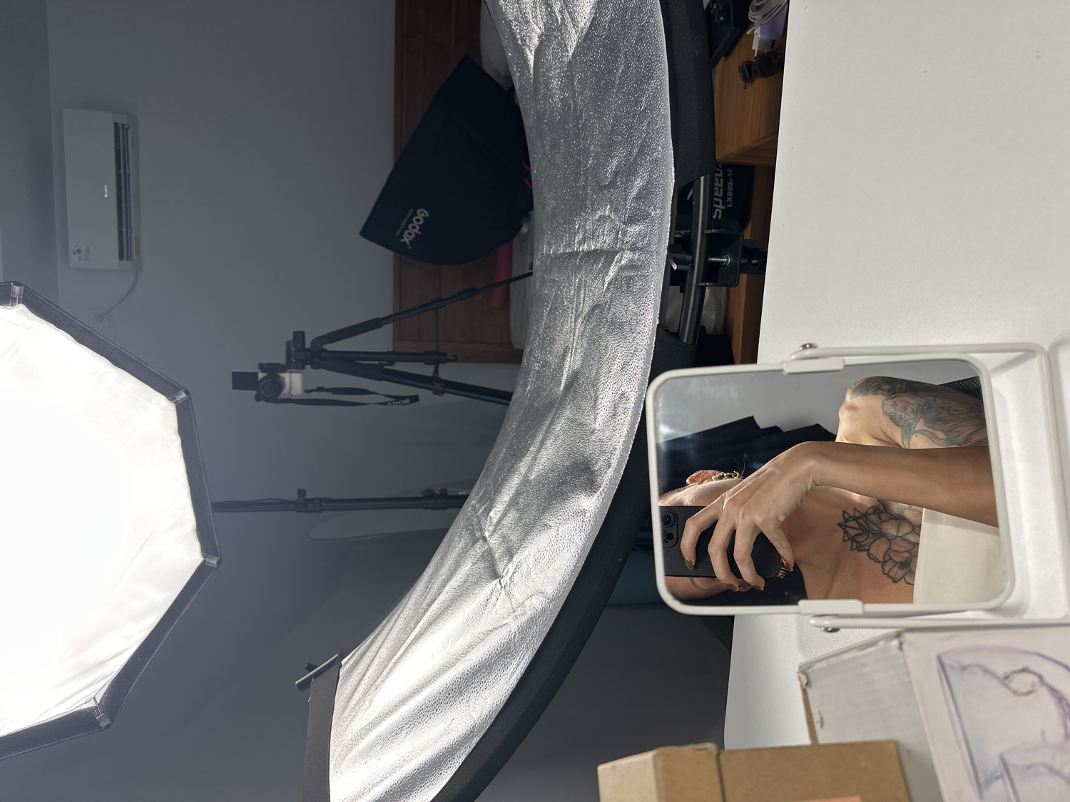An indoor scene, rotated 90 degrees clockwise, reveals a photography studio or a room equipped for photoshoots. The primary subject is a person's reflection captured in a small, rectangular mirror positioned on the right side of the frame. The reflection shows the person's upper torso, neck, and right arm, holding a smartphone to take the photograph. The person has several tattoos: a large floral design on the left shoulder/chest area, and another tattoo resembling an eagle or bird of prey on the upper right arm. Their right hand has painted fingernails and wears a golden ring.
The room is furnished with various photography equipment. A large, illuminated white octagonal softbox with a black rim is prominent on the left, serving as a light source. Another silver-lined strip softbox or reflector extends from the lower left. In the background, a small white camera, possibly a Sony (with "SONY" visible on its strap), is mounted on a black tripod. Another black light stand supports a black parabolic softbox or beauty dish, which prominently displays the "Godox" brand name.
Other details include a white air conditioning unit mounted on a light grey wall in the upper left background. A wooden shelf with items like sunglasses and small boxes is visible behind the mirror, bearing text like "Speed..." and "LX009-10". Below the mirror, brown cardboard boxes are stacked. The scene is brightly lit by the studio lights. The image captures a self-portrait being taken in what appears to be a functional studio environment in North Kuta, Indonesia.