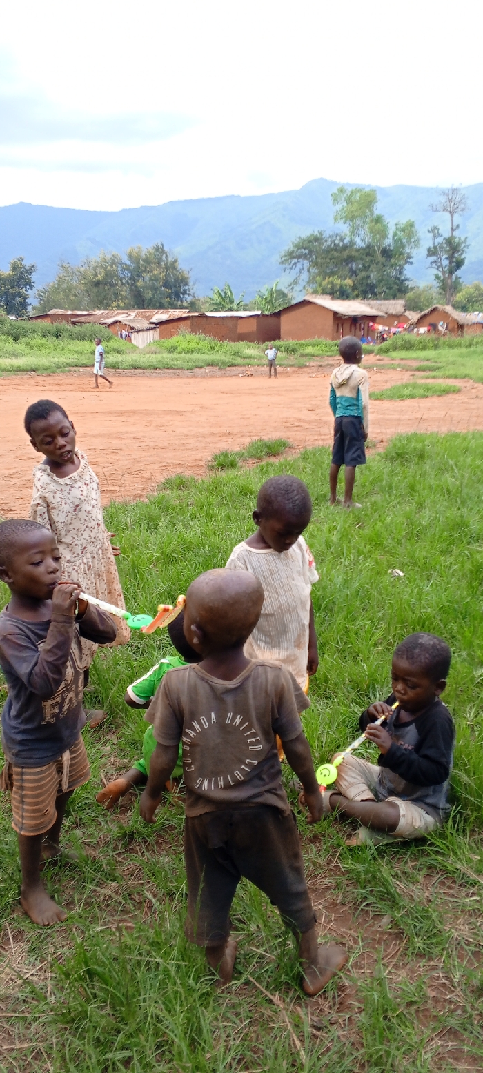 The image portrays a scene in Ngalula, Democratic Republic of the Congo. In the immediate foreground, approximately seven young children are gathered on sparse green grass. Two children are actively blowing party favor noise makers: one on the left wears a dark long-sleeve shirt and striped shorts, while another, seated on the right, wears a dark long-sleeve shirt and light pants. A third child, viewed from behind, wears a dark t-shirt with "RWANDA UNITED CLOTHING CO." inscribed on the back. Another child in a light floral dress looks toward the viewer, and a child in a light t-shirt looks downwards. All visible children in the foreground are barefoot and their clothing exhibits dirt and wear.
Behind this group, a wide, reddish-brown dirt area extends into the mid-ground. Two figures, identified as adult males or older boys, are positioned on this dirt path: one walks left in a white shirt and dark pants, and another stands further right in a light shirt and dark pants.
The background features multiple single-story buildings constructed with reddish-brown earth walls and rust-colored corrugated metal roofs. Green trees and foliage are interspersed among the structures, with laundry visible hanging outside one dwelling. Dominating the far background are large, verdant mountains beneath a pale blue, partially cloudy sky.