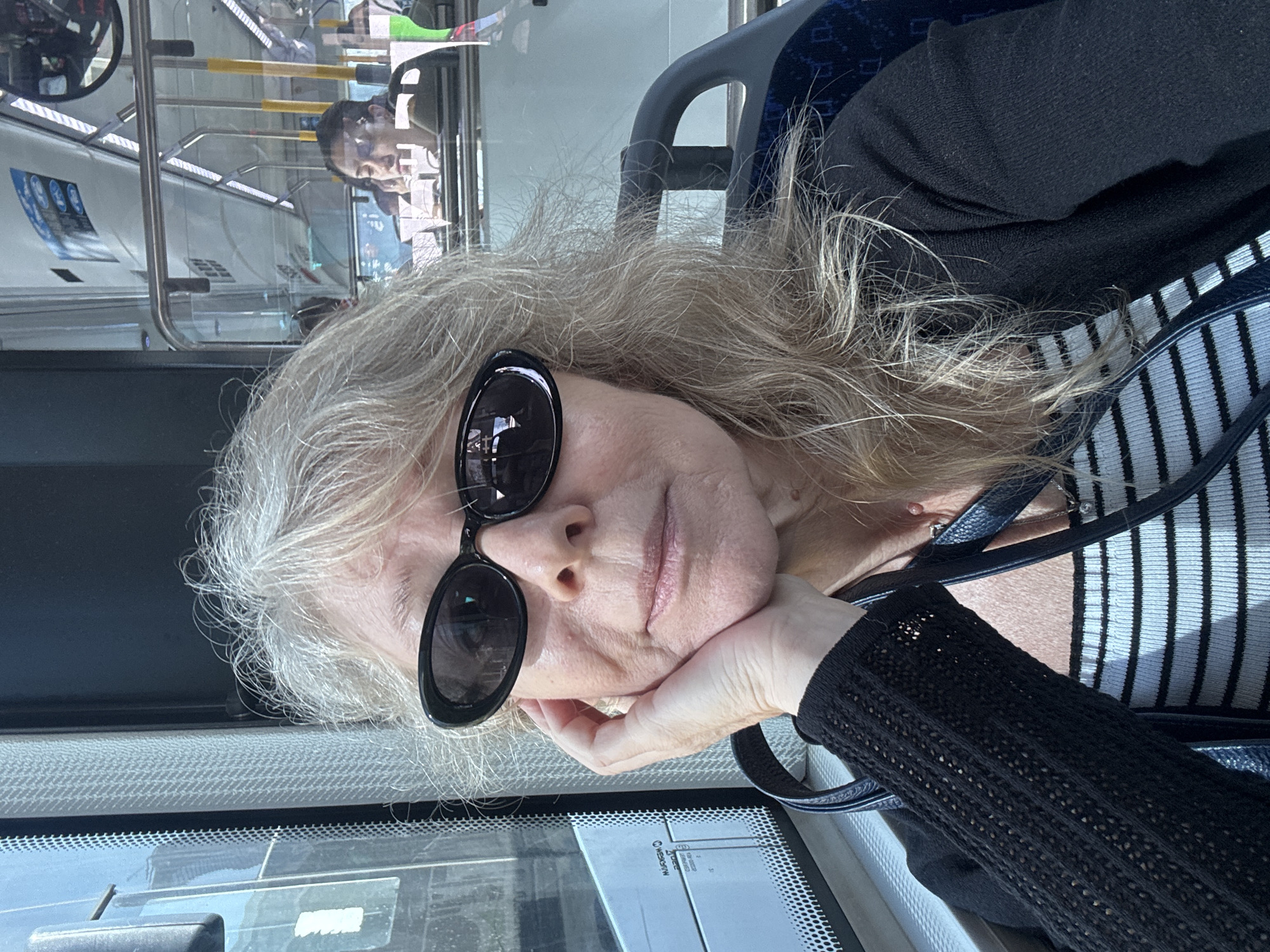 A woman with blonde hair and sunglasses is sitting on a bus, possibly in Tel Aviv, Israel. She is resting her chin on her hand and looking at the camera. The bus has blue seats and metal railings. In the background, other passengers can be seen. The woman is wearing a black cardigan over a striped shirt.