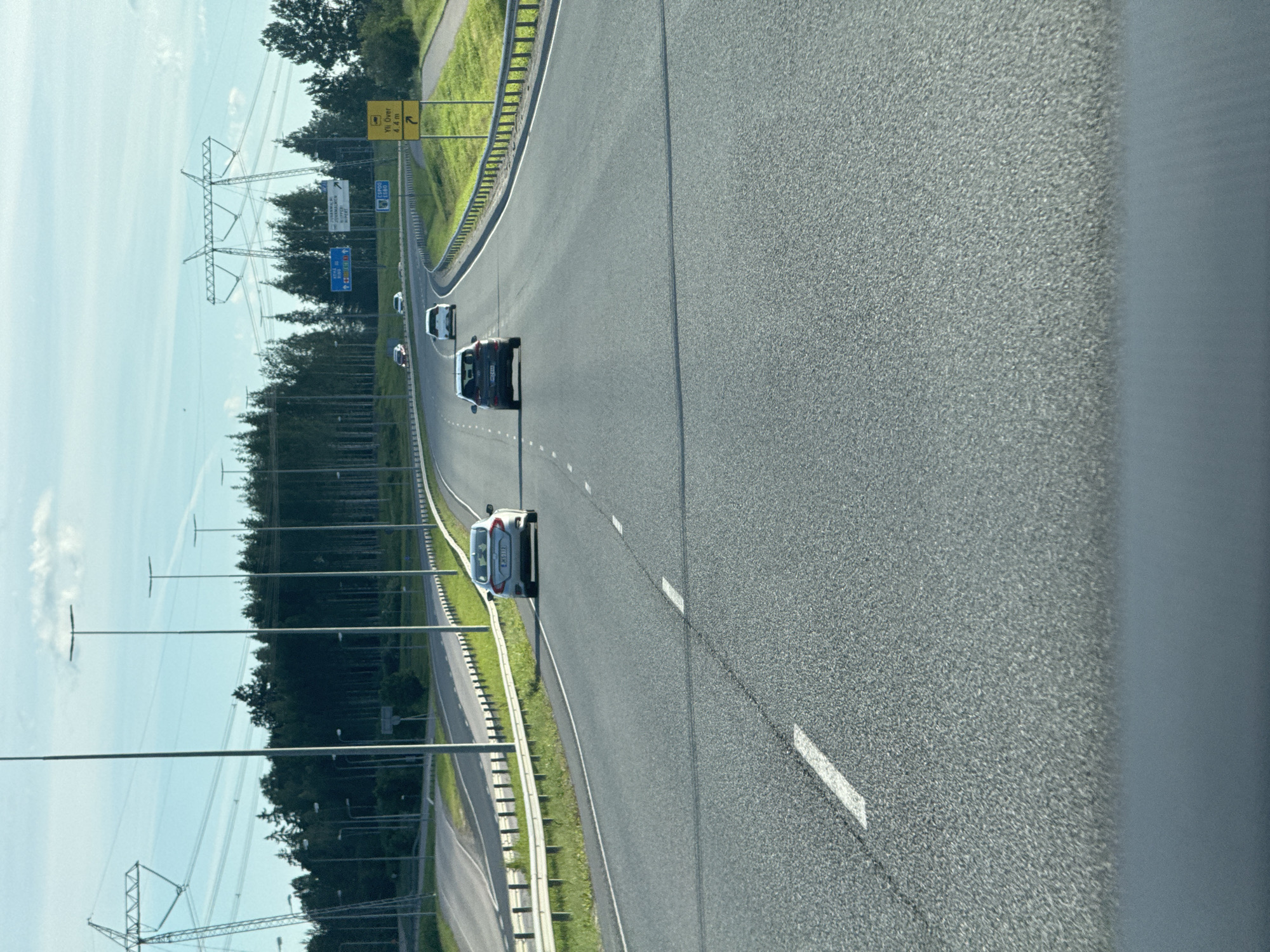 The image shows a highway curving to the right, with a consistent asphalt surface marked by white dashed lines. Several cars are traveling on the highway in the same direction. The scene is bordered on the left by a grassy embankment and a metal guardrail, and beyond that, a dense forest. Tall electrical pylons with numerous cables are visible against a bright, clear sky, suggesting an open area with power infrastructure. On the right side of the highway, the view is partially obscured by a blurry, bluish-gray area, possibly a vehicle window or a reflection.
There are no people clearly visible as subjects. The activity is vehicular traffic on a highway. Notable details include the sunny weather and daytime setting. The presence of highway signs indicates a road network. One sign visible has yellow text on a yellow background with a black arrow, and another sign appears to have blue text. The overall atmosphere is neutral and functional, depicting a common travel scene.
