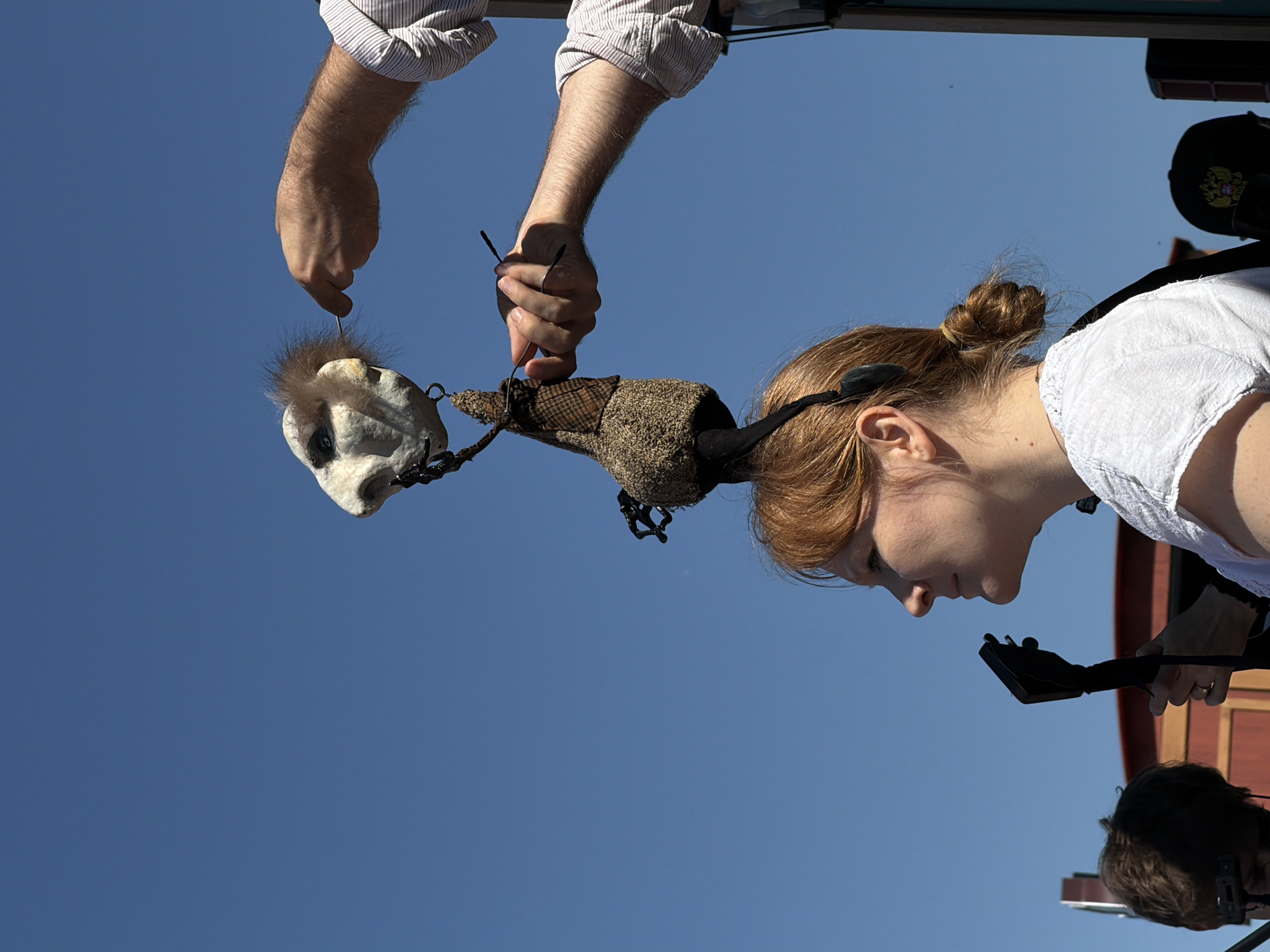An outdoor scene in Khimki, Russia, on a clear, sunny day shows a woman wearing an elaborate headpiece featuring a puppet, while a man manipulates the puppet with rods.
The central figure is a woman depicted in left profile, her reddish-blonde hair tied back in a bun. She wears a textured white short-sleeved top and a thin ring on her left ring finger. A black, head-mounted object is visible over her ear, and she holds a dark, guitar-neck-like object with her hands in the lower left portion of the frame. Above her head, a stylized puppet serves as part of her headwear. The puppet features an off-white, skull-like face with dark eye sockets and a prominent nose, topped with a tuft of brown fur. Its body is constructed from brownish, textured fabric with dark, chain-like embellishments.
Two male arms, belonging to one individual, are visible in the upper right. The left arm, wearing a light-colored striped shirt, extends to manipulate the puppet's lower body with a thin, dark rod. The right arm, similarly attired, holds another dark rod, touching the fur on the puppet's head.
The background consists of a clear blue sky. Partially visible on the far left is the back of another person's head, possibly wearing an electronic device. To the woman's left, a blurred reddish-brown structure, possibly part of a stage or building, is present. On the far right, a dark structure and the top of a black baseball cap with a golden emblem are discernible.