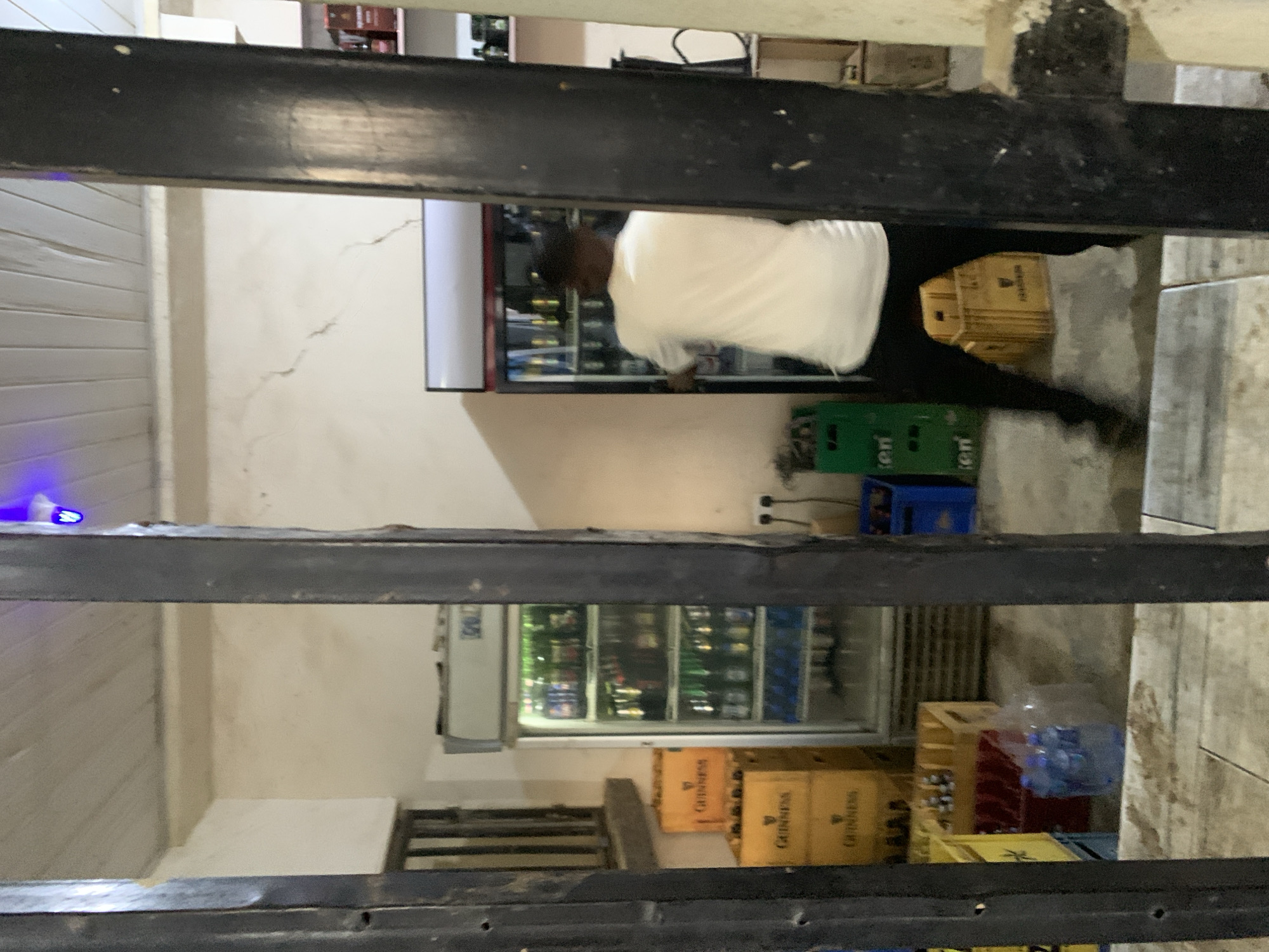 The image depicts the interior of a commercial establishment, likely a beverage store or bar, located in Kabong, Nigeria. The photograph is taken through dark, vertical bars, partially obscuring the view.
In the center-right, a person is visible from the back, wearing a white t-shirt and dark trousers, actively engaging with a large, glass-fronted commercial refrigerator. This refrigerator, positioned on the right side of the space, contains multiple shelves of bottled beverages. To the left of the person, another similar large, glass-fronted refrigerator is stocked with various bottled drinks. Stacked beside the left refrigerator are several yellow wooden crates, prominently branded "GUINNESS." More "GUINNESS" branded crates are present on the floor further left.
The walls are painted an off-white or cream color. The wall behind the left refrigerator exhibits multiple vertical cracks. A white paneled ceiling is overhead, with a blue light fixture visible near the top center. The floor is a concrete or tiled surface, showing some staining and wear.
Additional details include a wooden railing on the far left, suggesting a raised section or staircase. On the floor in the foreground, a transparent plastic package of bottled water is present alongside other colored plastic crates (green, blue, red) containing bottles. On the far right, shelving against the wall displays various bottles, likely spirits. Electrical outlets are visible on the right wall.