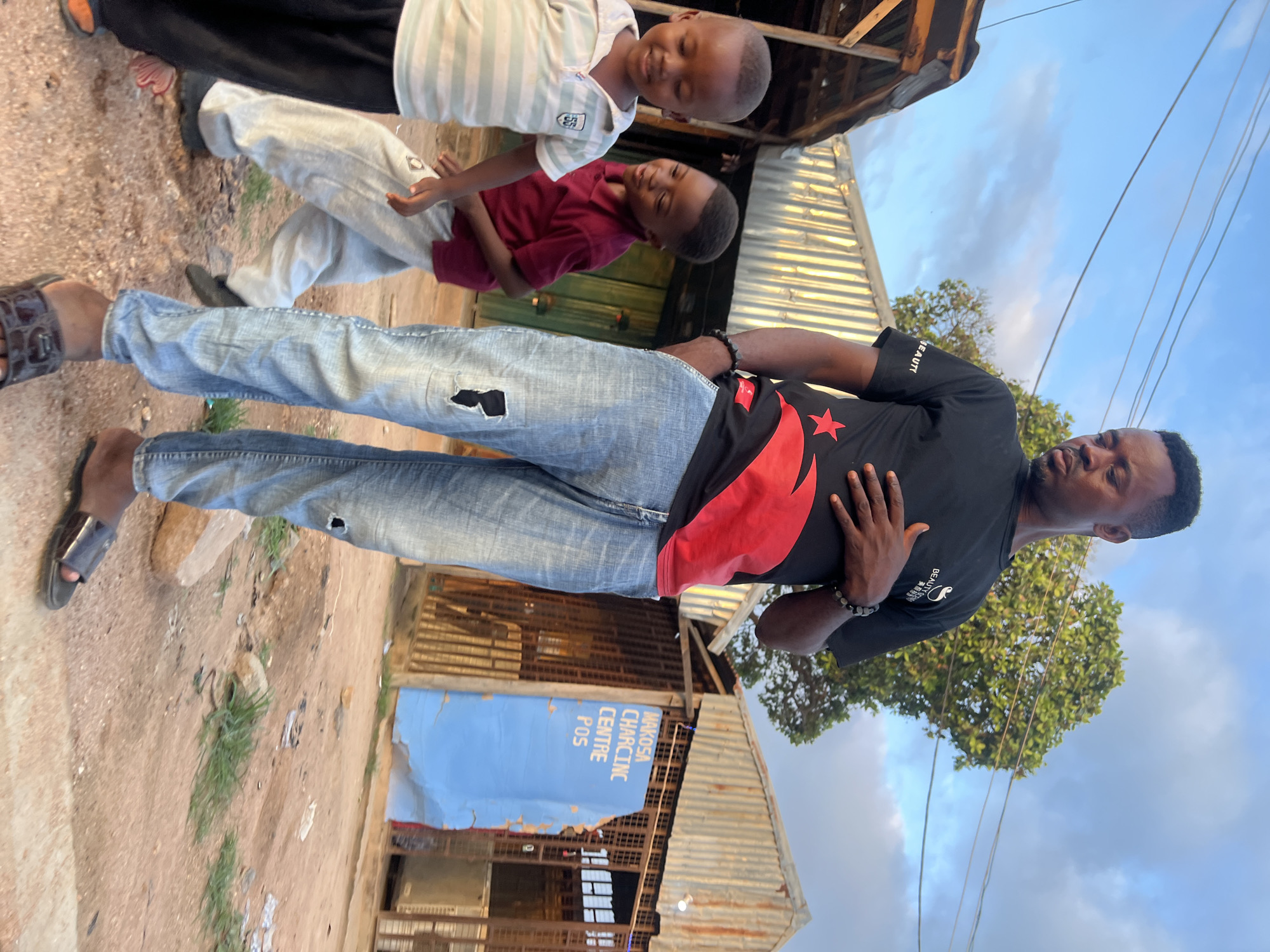 The image, correctly oriented (rotated 90 degrees counter-clockwise from its current display), depicts an outdoor scene in the city of Howayi, Nigeria.
In the foreground, an adult male stands centrally, wearing a black short-sleeved t-shirt with a red star and a wing-like design on the left chest. He wears faded blue ripped jeans and dark sandals with brown straps. A beaded bracelet is visible on his left wrist, and his left hand rests on his stomach. He has short dark hair and a light beard, with his gaze directed downwards. To the far left, partial view of another person's legs and feet, clad in blue jeans and dark sandals, is visible.
In the mid-ground, two young boys are positioned to the right of the central adult. The boy closer to the viewer wears a maroon short-sleeved shirt and light-colored pants, with dark shoes, looking towards the left with a slight smile. The second boy, to his right, wears a light-colored, possibly striped, short-sleeved polo shirt and dark pants, also looking left with a slight smile.
The ground beneath the subjects is unpaved, consisting of packed dirt with sparse patches of green grass and scattered debris.
The background features various structures. A prominent building displays a large blue sign with white text that reads "MAKOSA CHARGING CENTRE POS," indicating a charging station and point-of-sale service. Other buildings appear constructed from corrugated metal sheets and wooden frames. To the left, a large tree with dense green foliage is visible. The upper portion of the scene is dominated by a light blue sky with scattered white clouds, crossed by overhead utility wires.