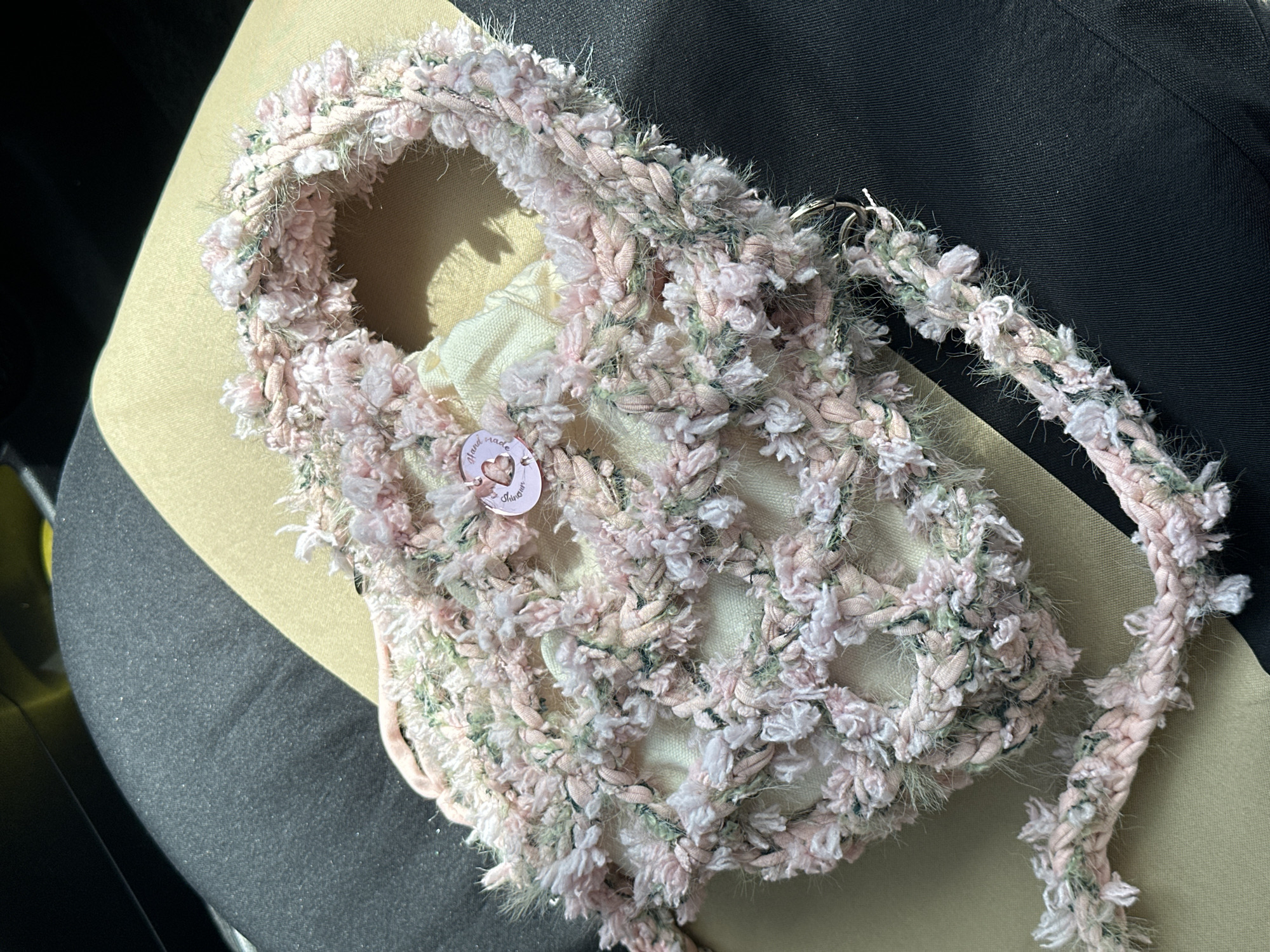 This is a close-up, high-angle, eye-level shot of a handmade crocheted bag resting on a car seat in a city. The bag is primarily pink with some green and white accents, featuring a looped, open-weave pattern. A small pink button with a heart and text is attached to the front of the bag. The car seat appears to be upholstered in a dark grey fabric with a lighter tan fabric underneath. The background is blurred, but suggests the interior of a vehicle, possibly with sunlight filtering through. The image does not contain any specific identifiable landmarks or features that would place it in Ko Samui District, Thailand.
