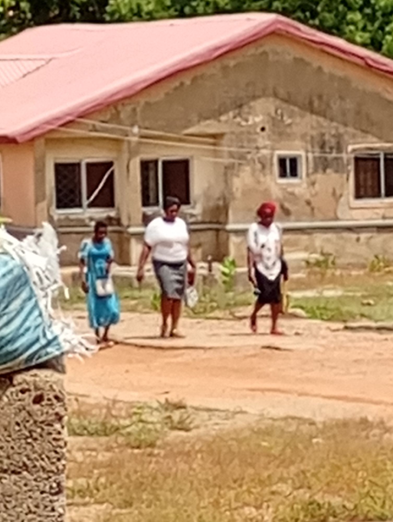 The image depicts three women walking on a dirt path in front of a one-story building with a pink roof and weathered, beige walls. The woman on the left is wearing a blue dress. The woman in the center is wearing a white top and a gray skirt. The woman on the right has a red head covering and is wearing a white top and dark skirt. The location is in the city of Unknown, Unknown.