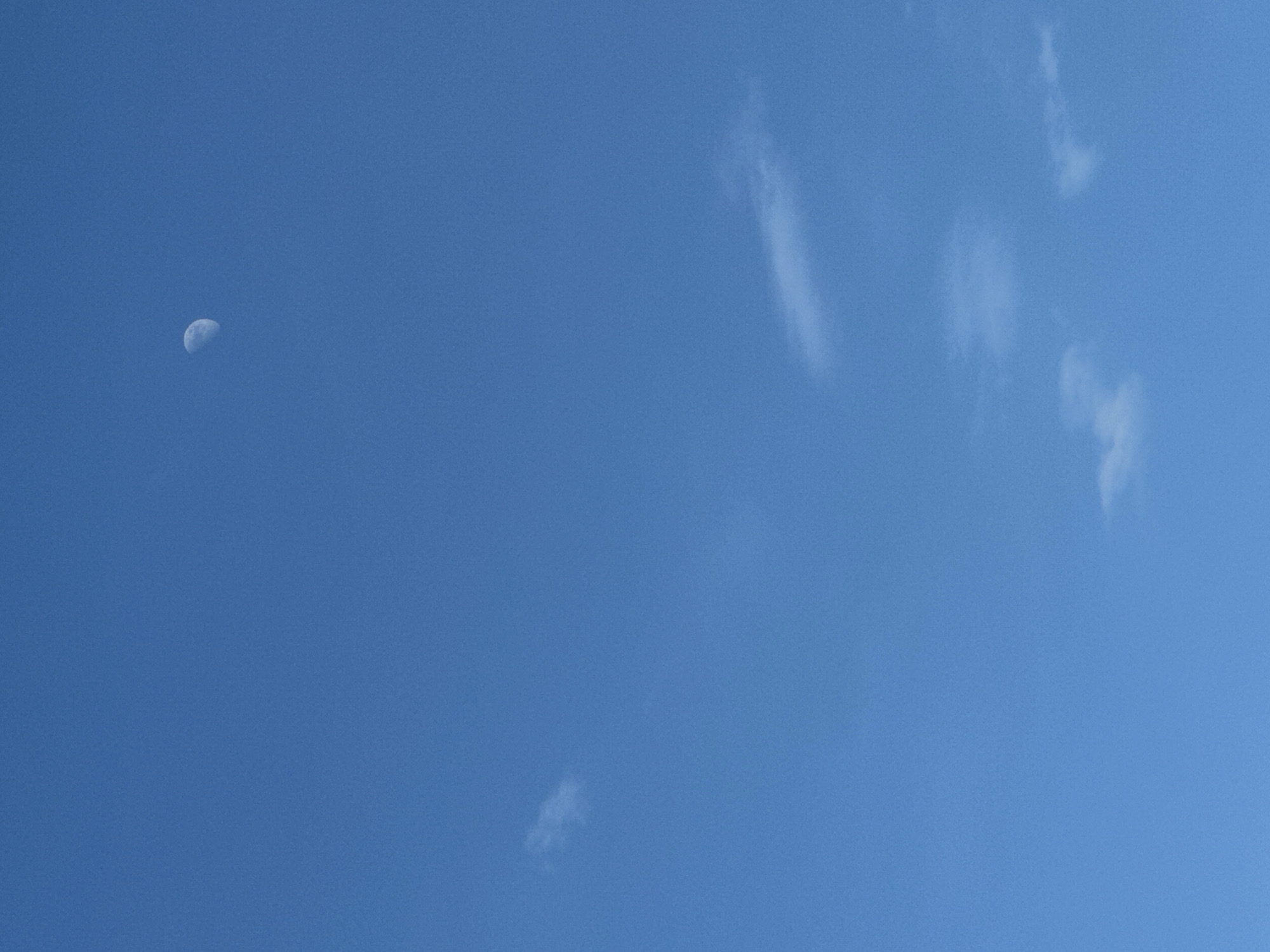 The image shows a daytime sky with a waning crescent moon visible in the upper left portion. Thin, wispy clouds are scattered across the sky, mainly in the right half of the frame. The sky is a clear blue. The location in the city of Chaikovsky, Russia is unknown. There are no people, specific subjects, or objects other than those mentioned. No actions or interactions are depicted, and the environment is limited to the sky.