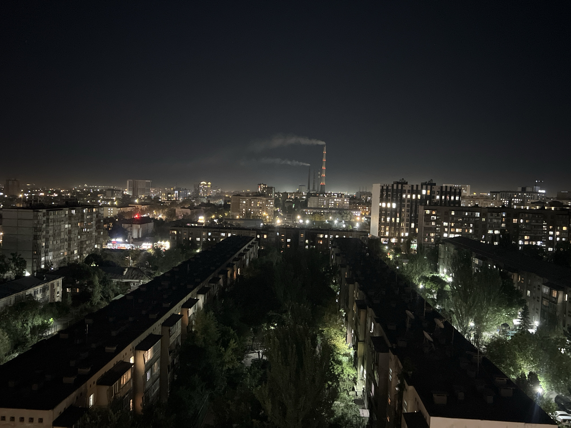 This is a night aerial view of a dense urban residential area, likely Bishkek City, Kyrgyzstan. The scene is dominated by apartment buildings of varying heights, their windows illuminated with warm light, suggesting occupancy. The buildings are arranged in a somewhat linear fashion, with courtyards and green spaces interspersed between them. Trees, some with dense foliage, are visible in these courtyards and along the streets, their leaves catching the ambient light.
In the distance, a prominent industrial smokestack is visible, emitting a plume of white smoke, tinged orange by its internal lights. The sky above is dark and overcast, obscuring most stars but allowing the city lights to create a haze. Other distant buildings and lights suggest a larger urban expanse beyond the immediate foreground. The overall impression is one of a typical, lived-in urban environment at night. There are no people or specific actions immediately apparent in the image, focusing instead on the architectural and environmental elements of the city.